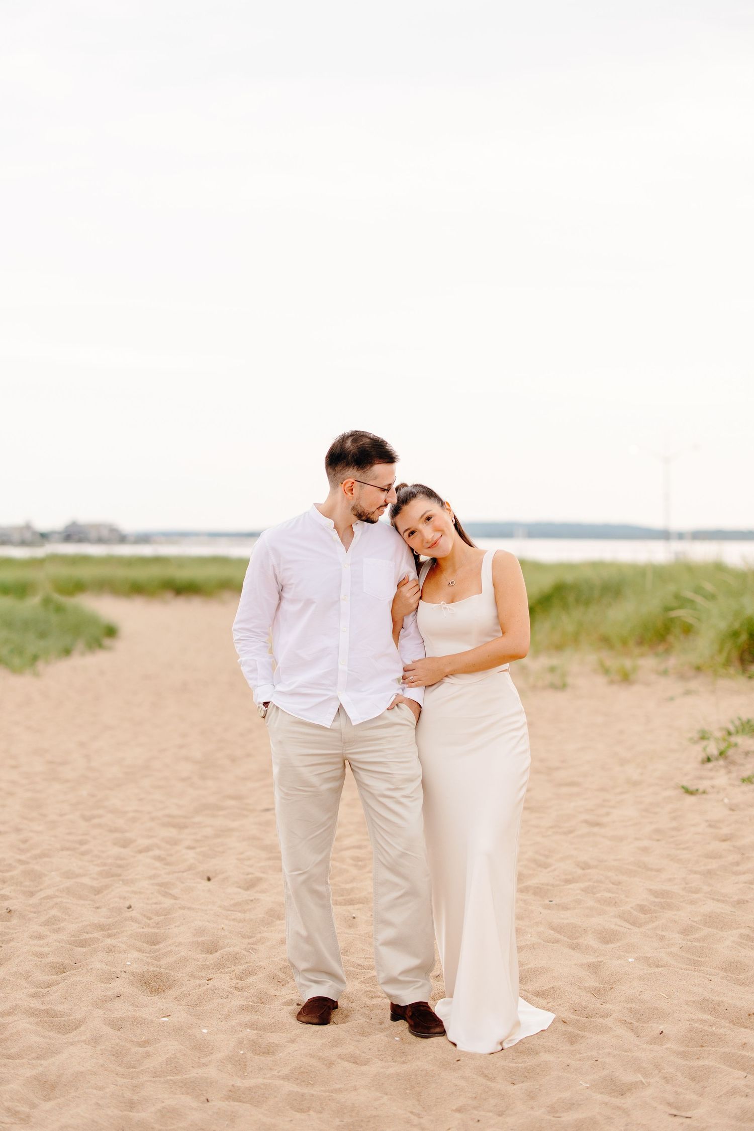 Couple embracing on sandy beach, woman in white dress leaning on man in white shirt, coastal grass background.
