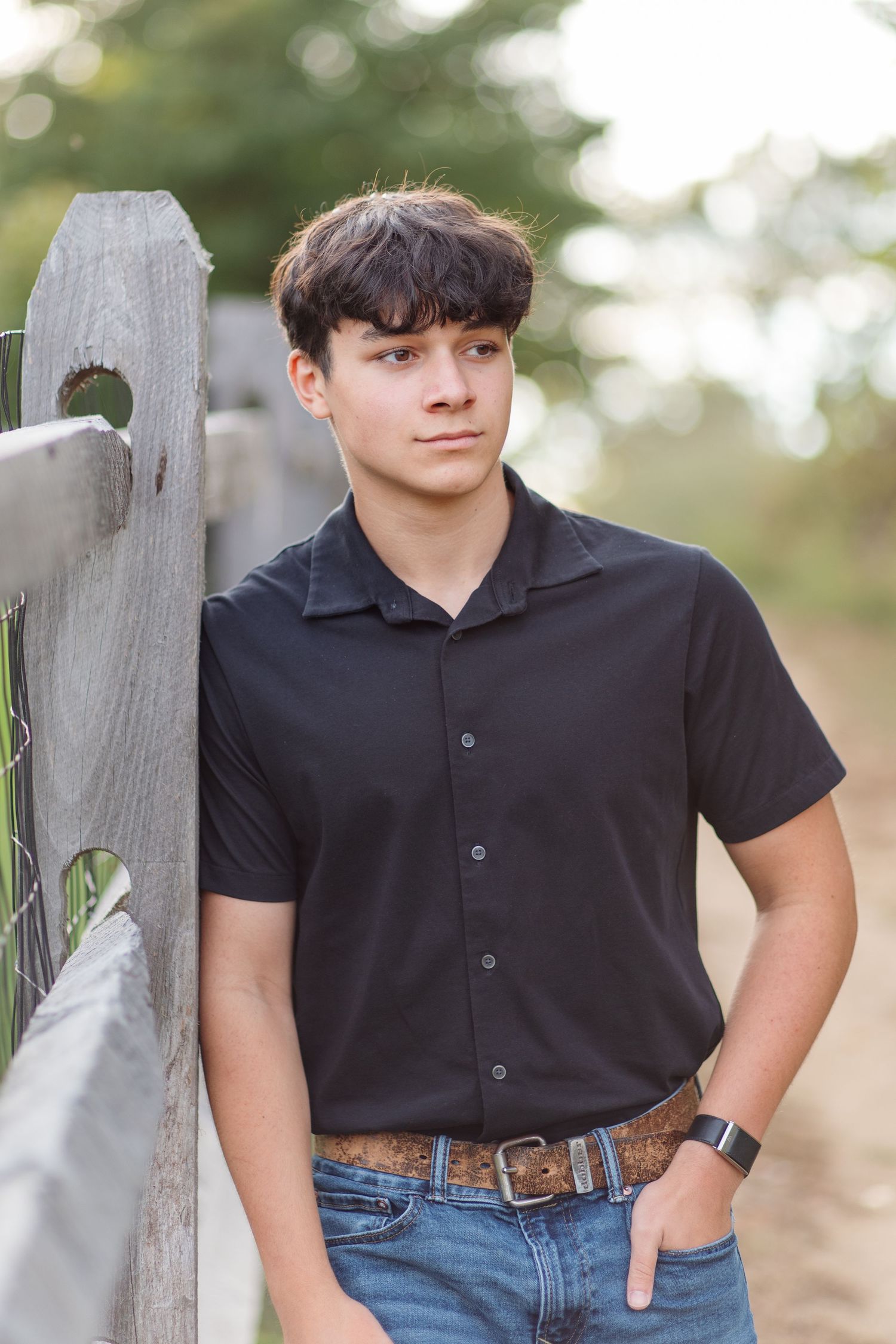 Teen boy in black shirt and jeans leaning against wooden fence on rural path.