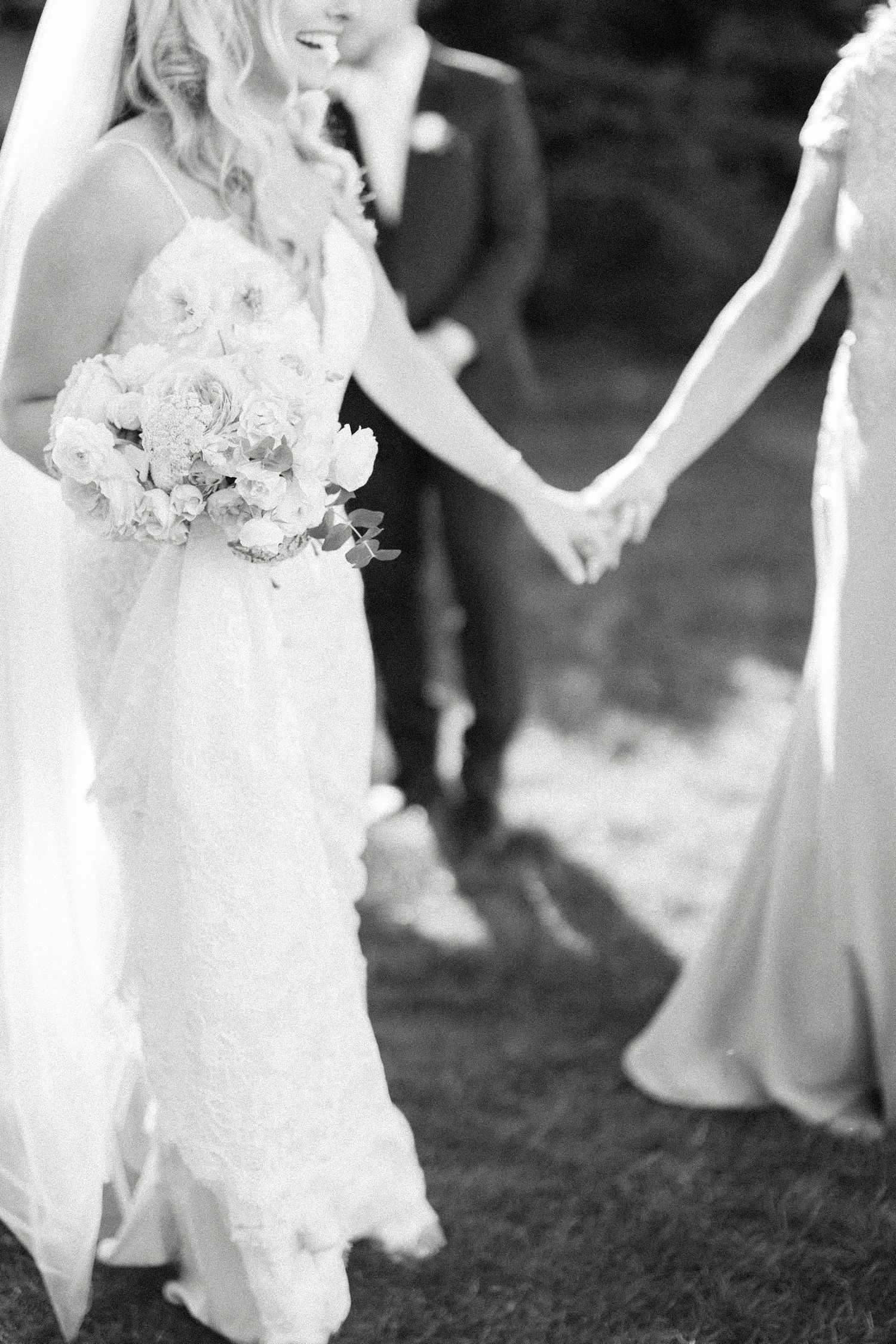 Black and white photo of bride holding a floral bouquet while holding hands with wedding party.