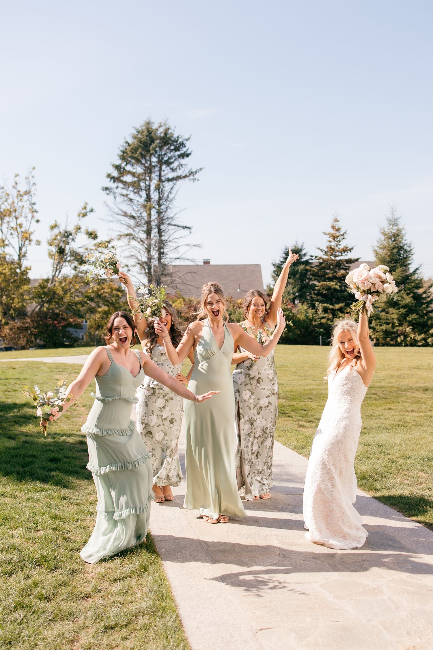 Bride and bridesmaids in sage green dresses celebrate joyfully on a sunny outdoor pathway.