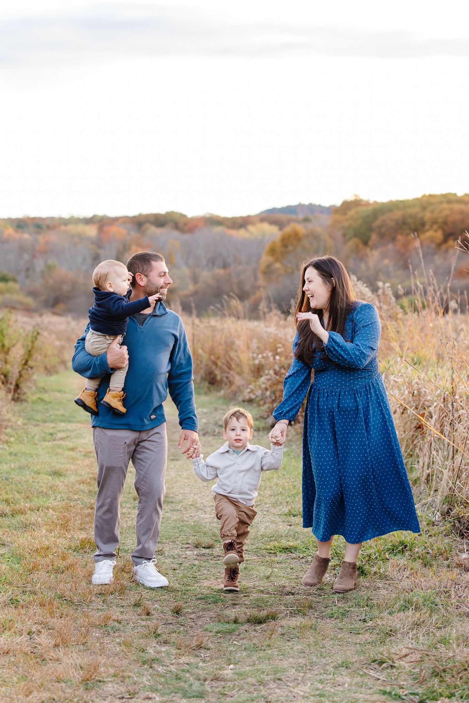 Joyful family of four walking through autumn field, father carrying toddler while son runs ahead.