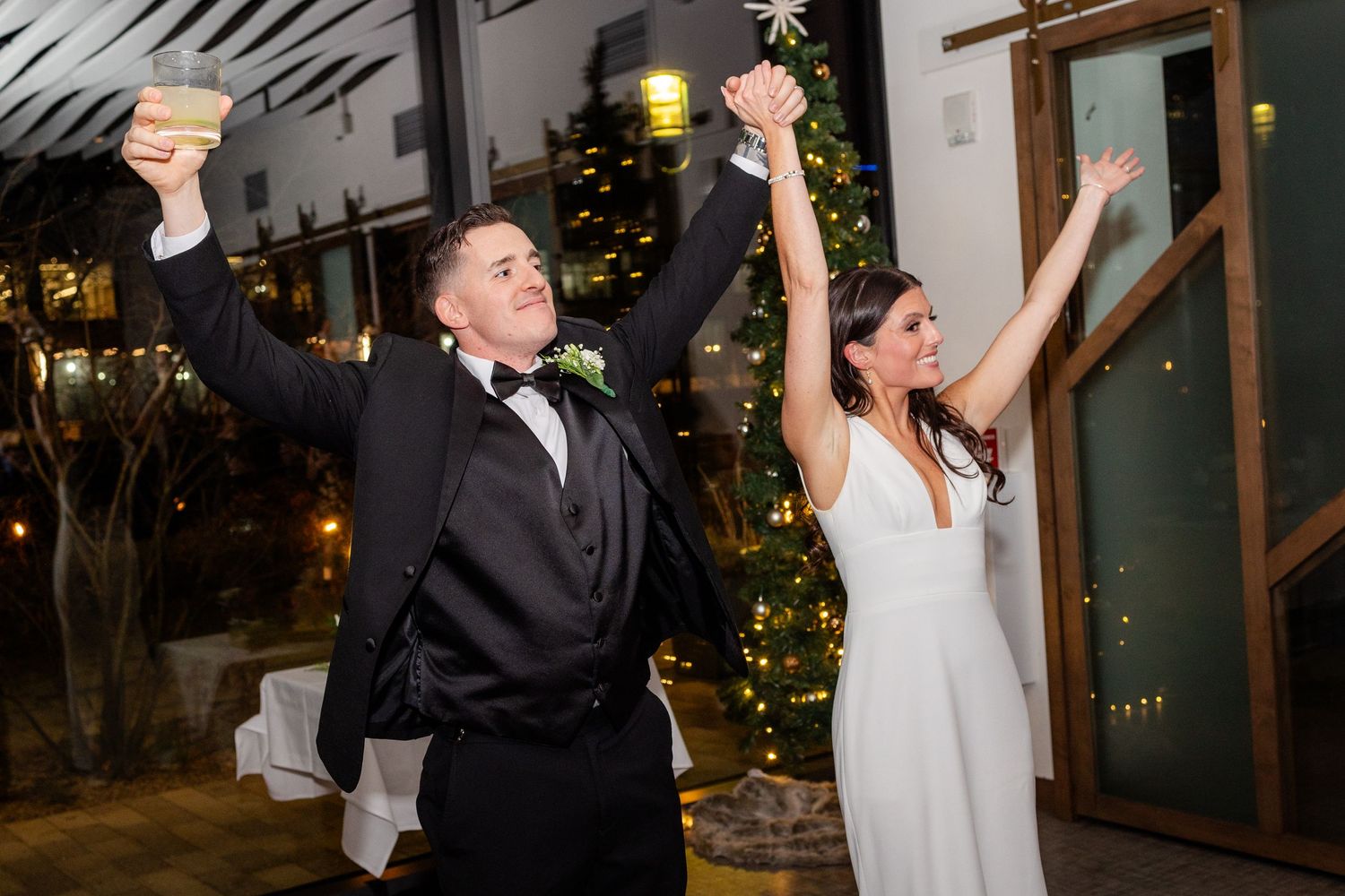 Bride and groom raise arms in celebration during festive wedding reception entrance near Christmas tree.