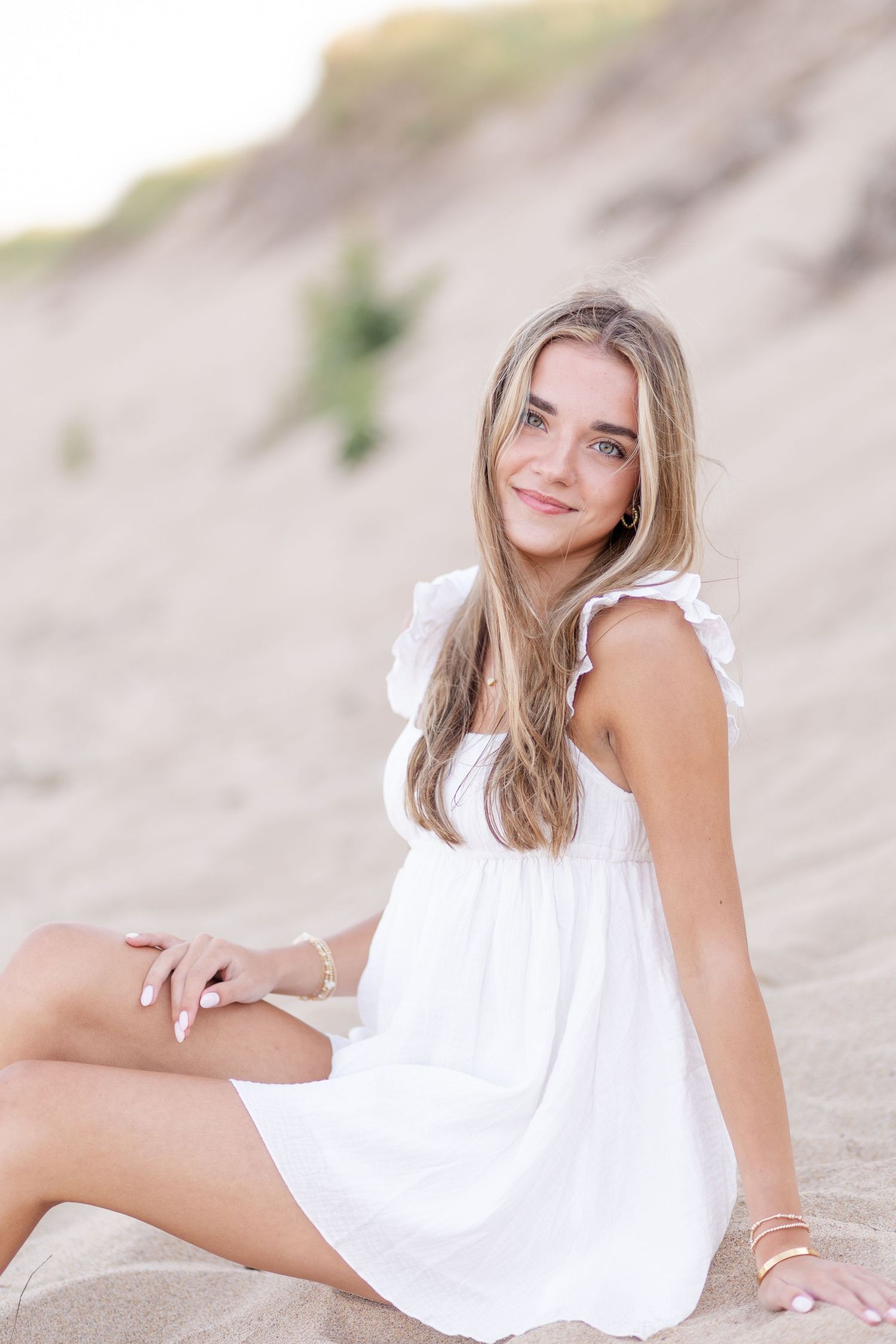 Blonde girl in white ruffled dress sitting on sandy dunes smiling at camera.