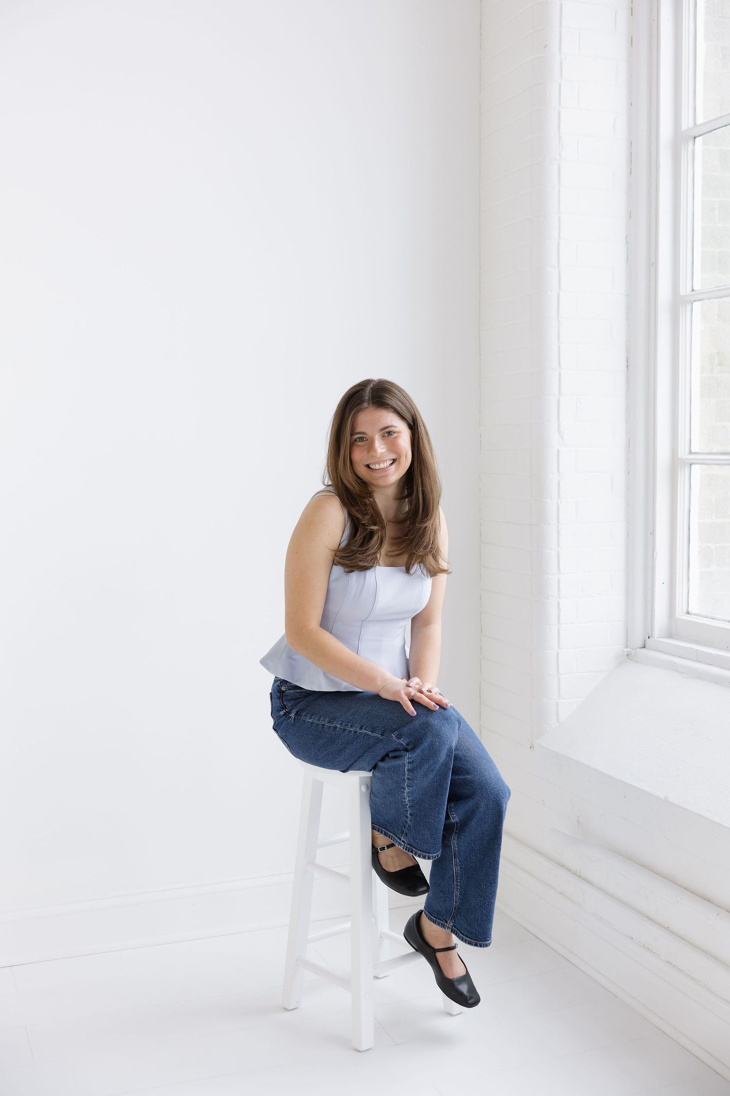 Smiling young woman in white top and jeans sitting on stool in bright white studio.
