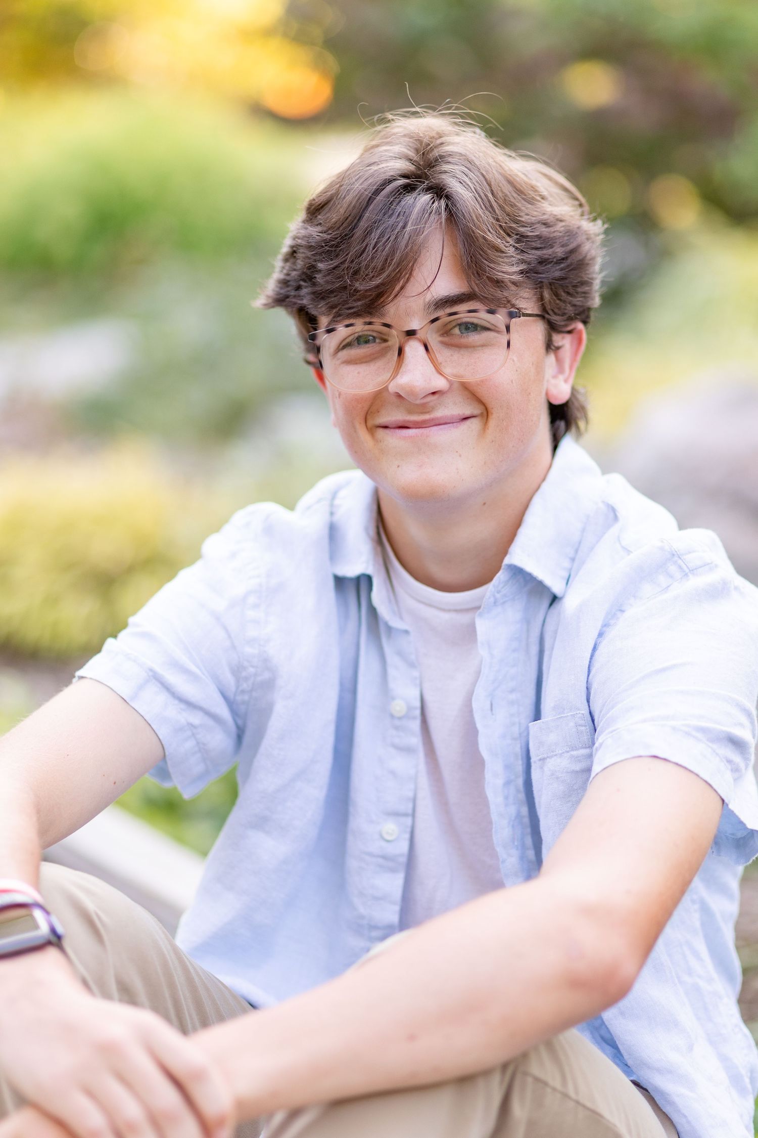Smiling teenage boy with glasses in light blue shirt sitting outdoors in a garden setting.