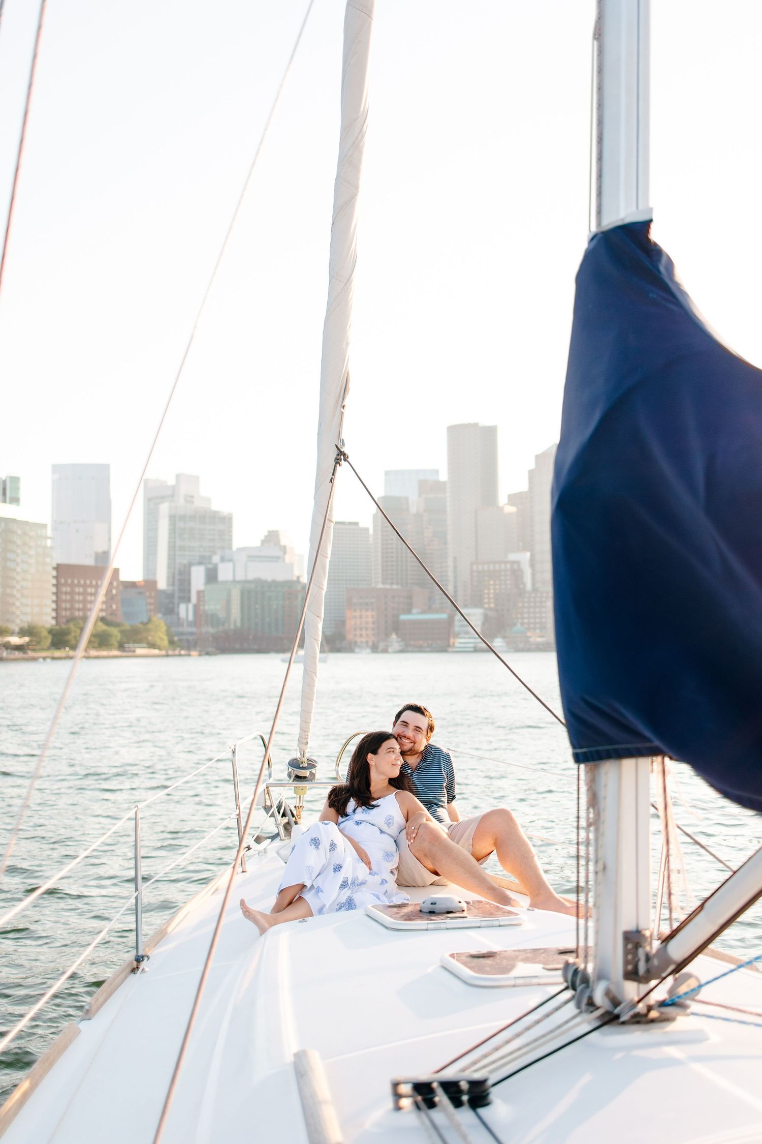 Couple relaxing on sailboat deck with city skyline backdrop on sunny day.