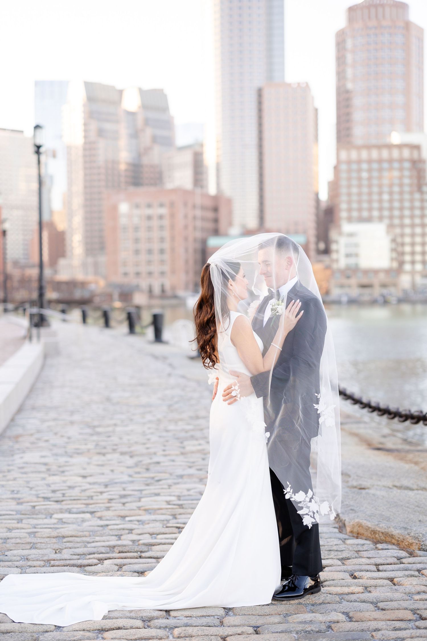 Bride and groom share romantic moment under veil on cobblestone waterfront with Boston city skyline backdrop.