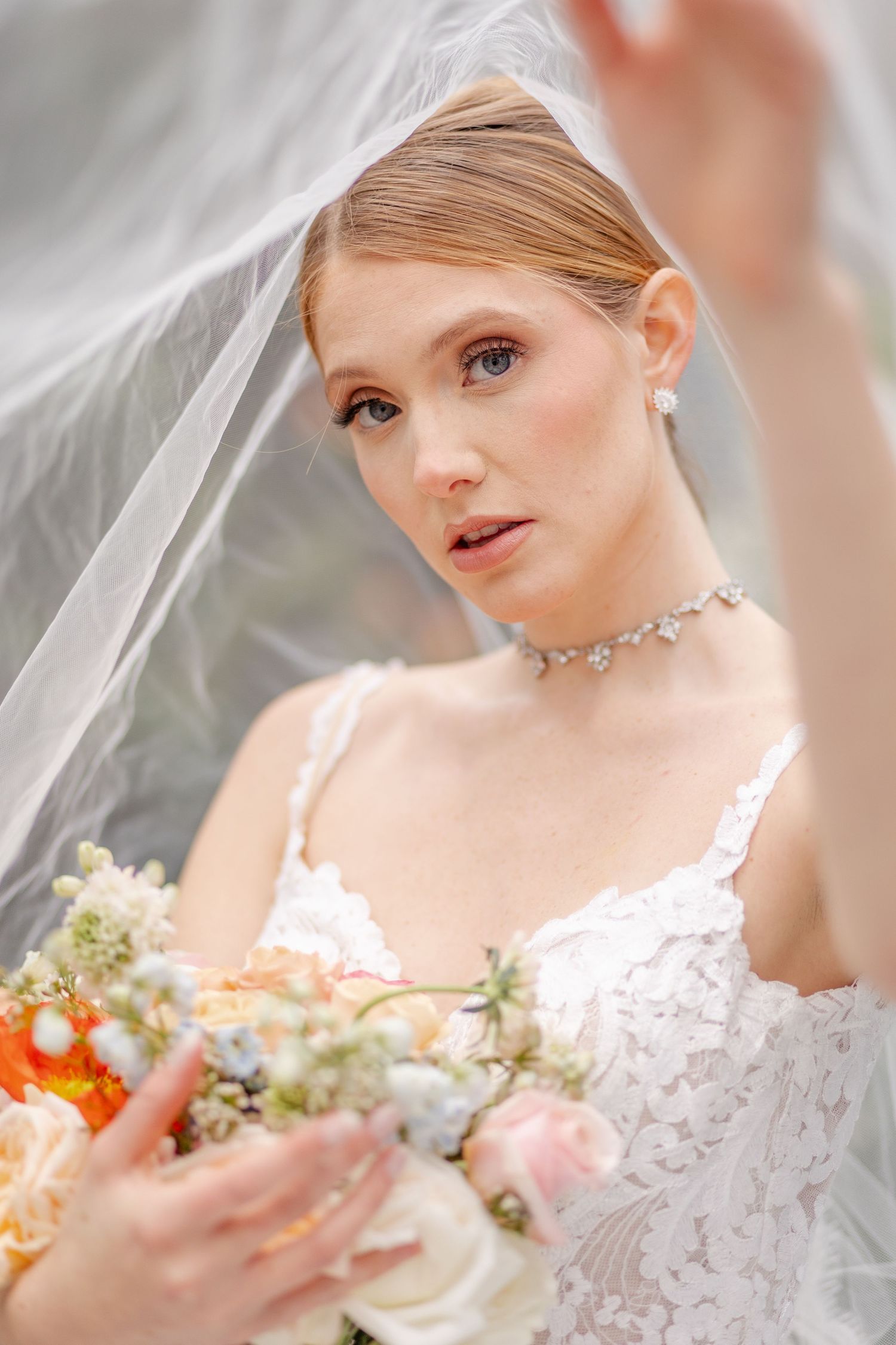 Bride in lace gown lifts veil while holding colorful bouquet with diamond choker necklace.