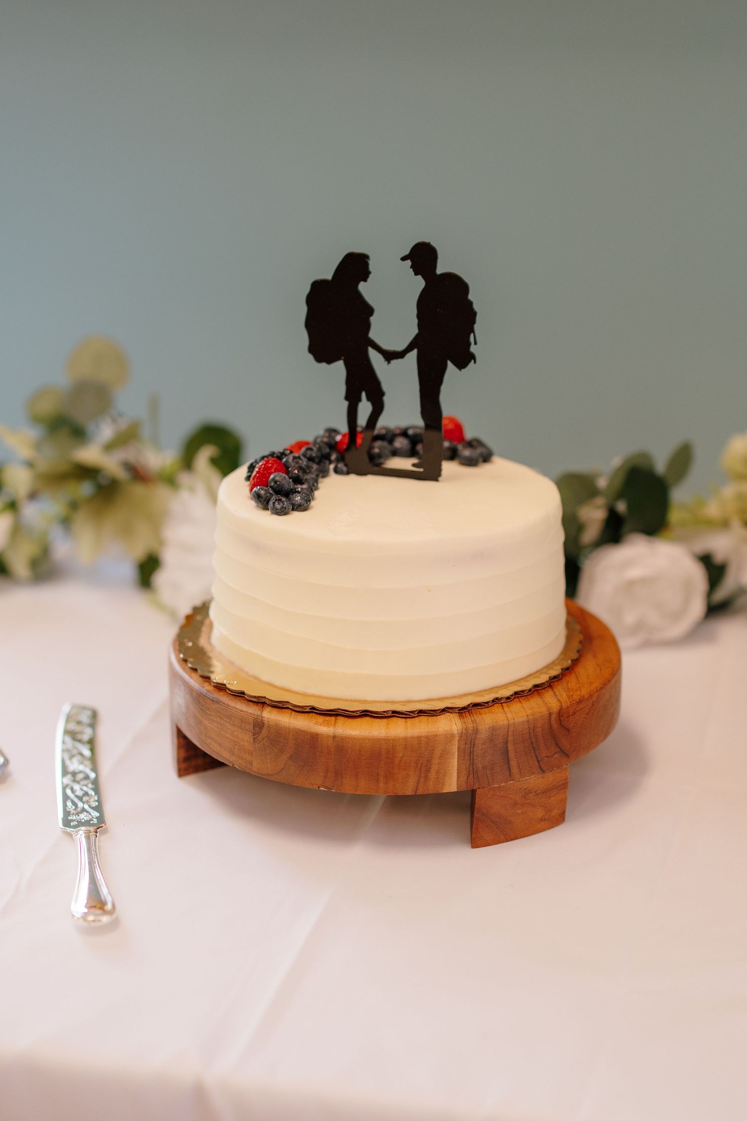 Elegant white wedding cake topped with hiking couple silhouette, decorated with fresh berries on wooden stand.