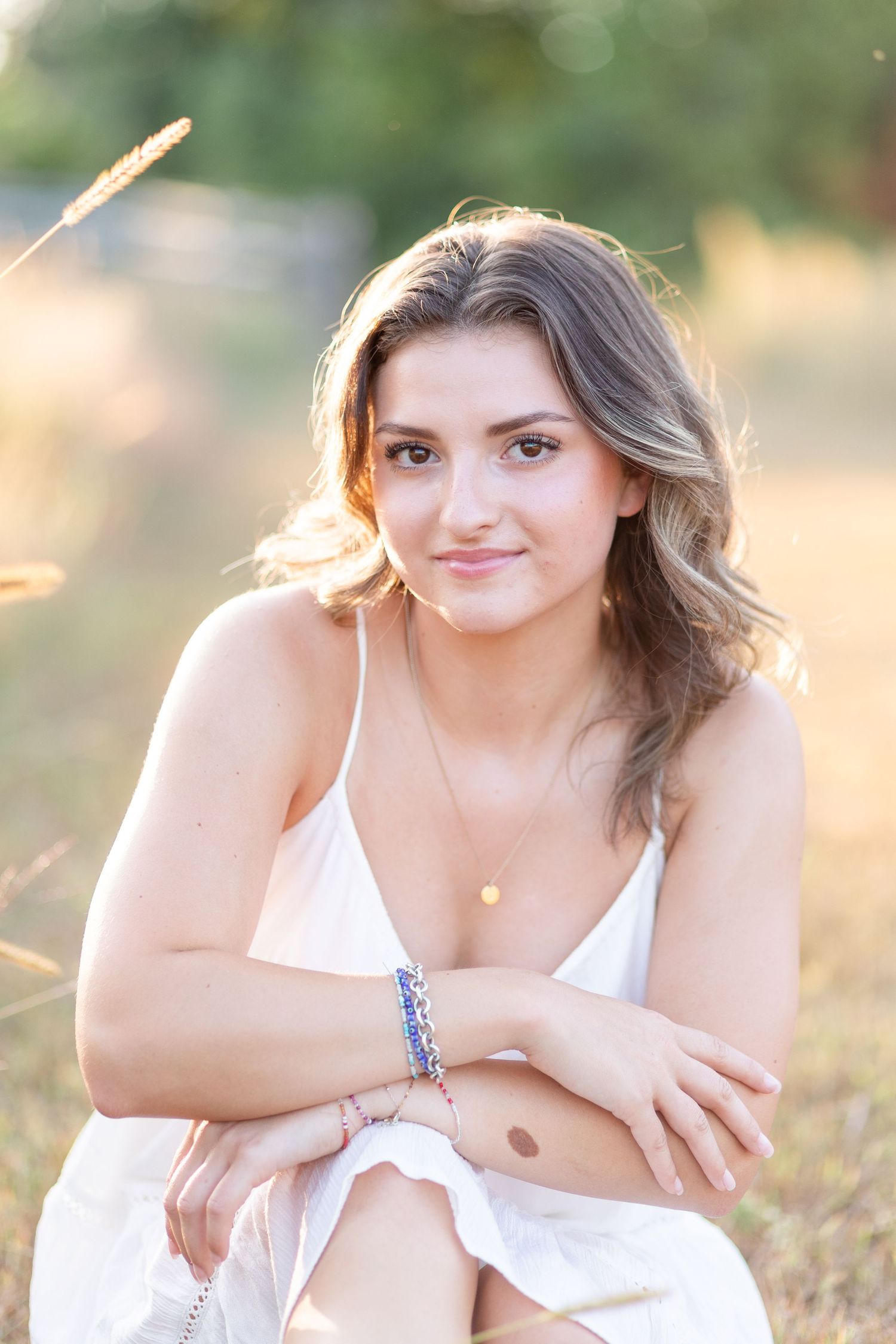 Young brunette woman in white dress sitting in golden field, wearing colorful bracelets, smiling softly.