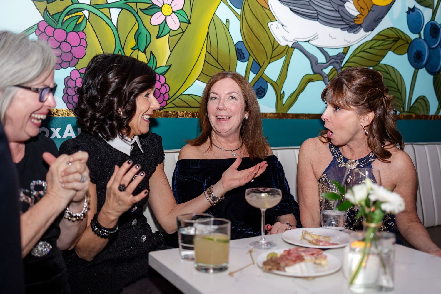 Four women laughing and conversing at wedding reception table with floral mural backdrop.