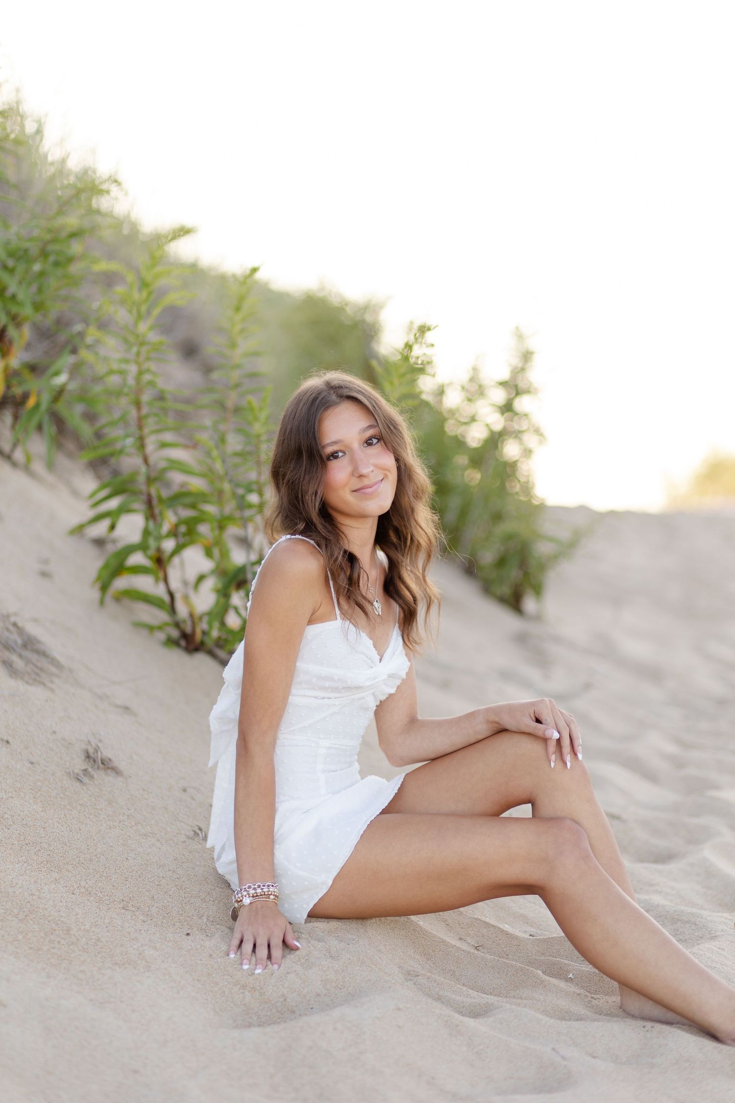 Young woman in white sundress sitting on sand dunes surrounded by green plants.