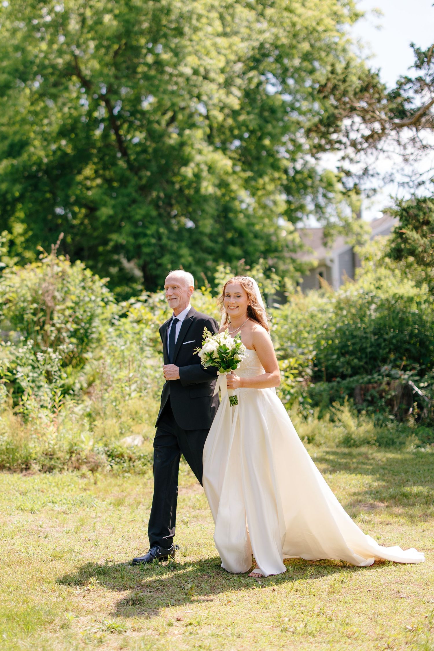 Bride in elegant white gown walking with father through lush green garden on wedding day.