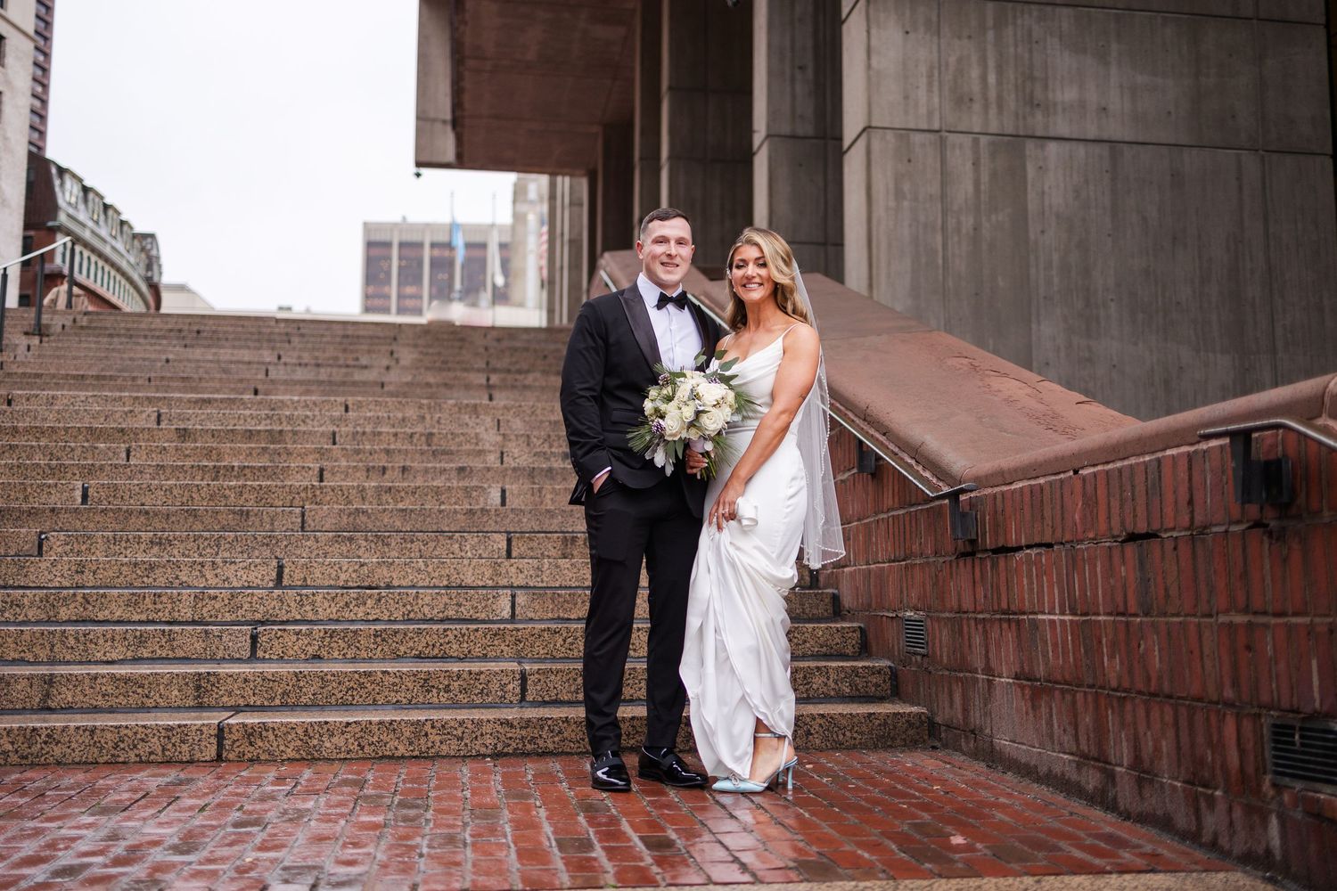 Elegant bride and groom posing on rain-soaked steps outside a grand building on their wedding day.