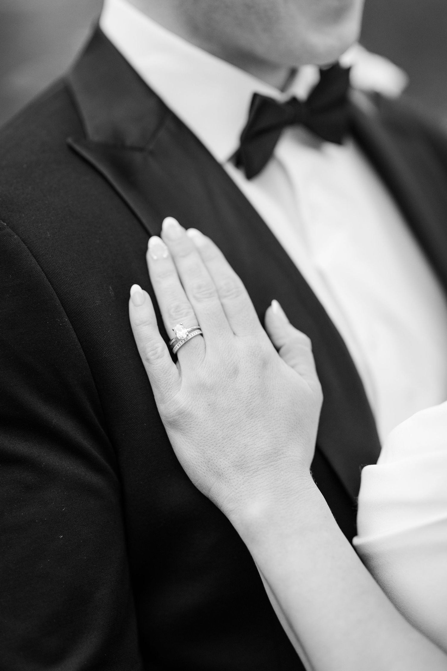 Black and white close-up of bride's hand with wedding ring resting on groom's tuxedo chest.