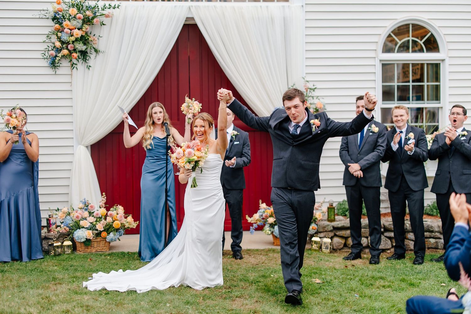 Bride and groom celebrate with raised hands at outdoor barn wedding ceremony with wedding party.