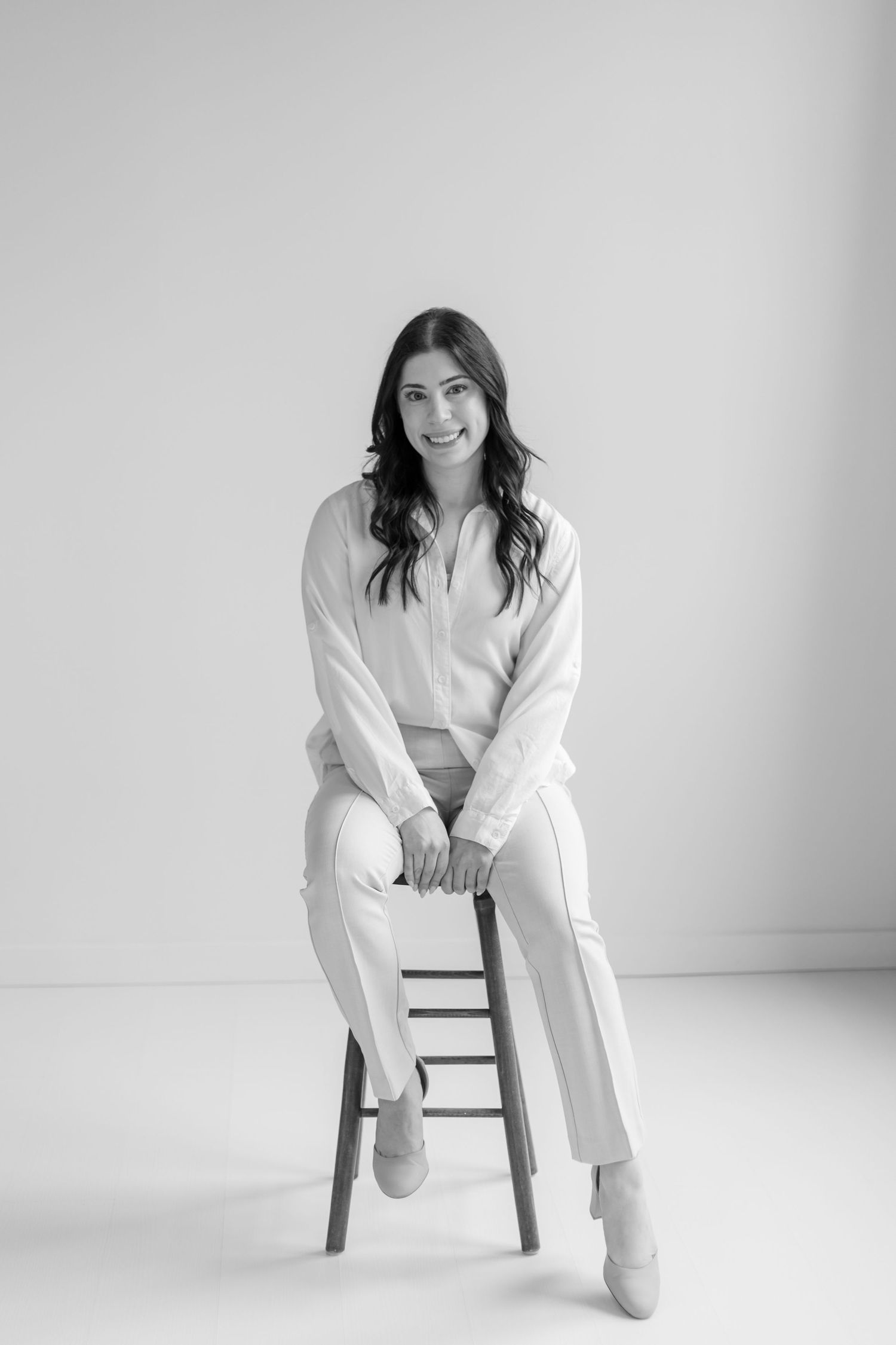 Elegant brunette woman in white blouse and trousers sitting on a stool, smiling in a black and white portrait.