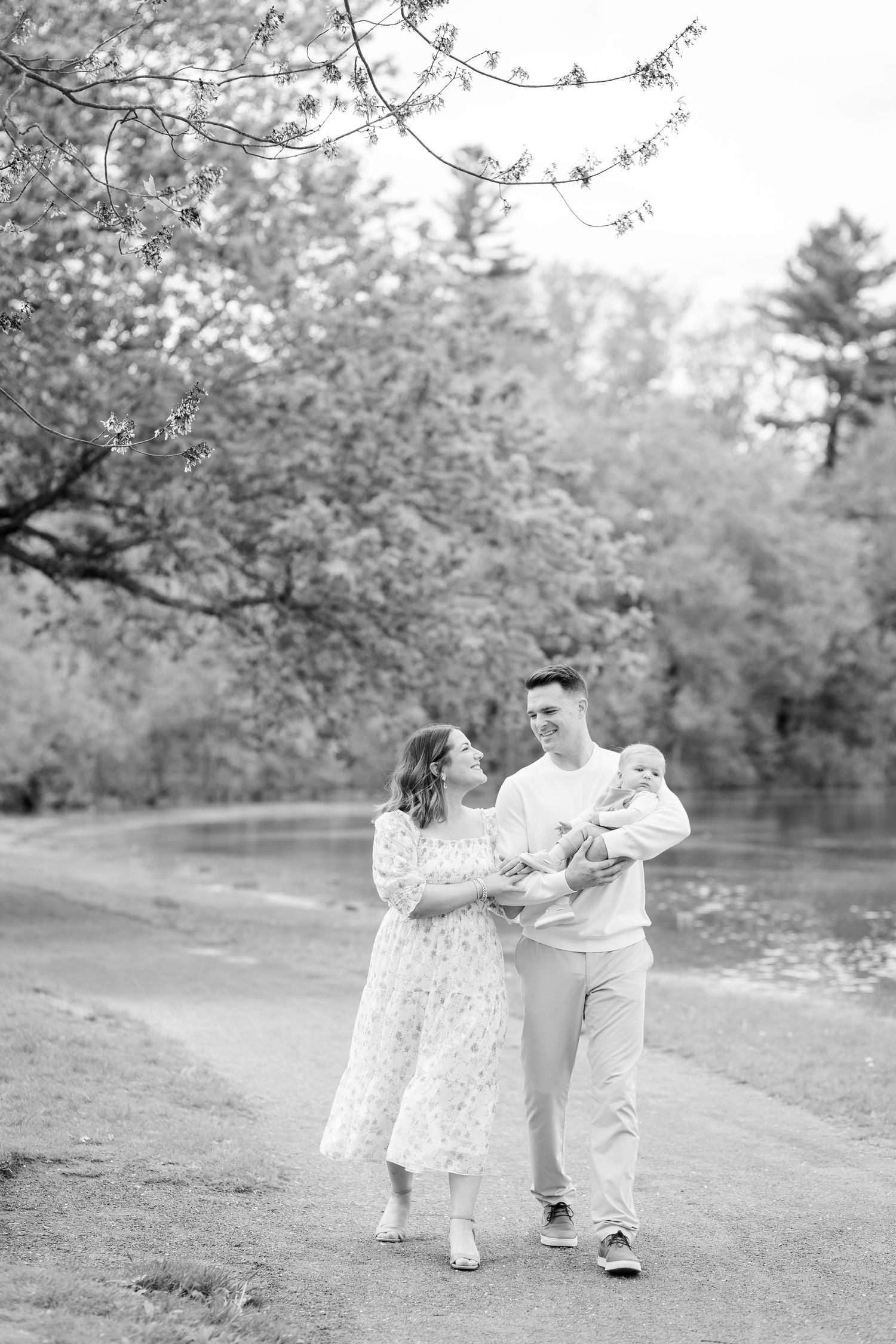 A black and white photo of a young couple walking with their newborn baby along a tree-lined path near a pond.