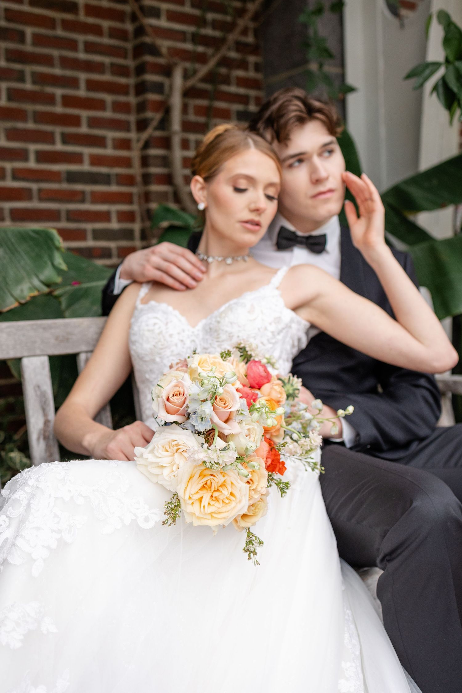 Bride and groom sit on bench, she holds peach and coral bouquet.