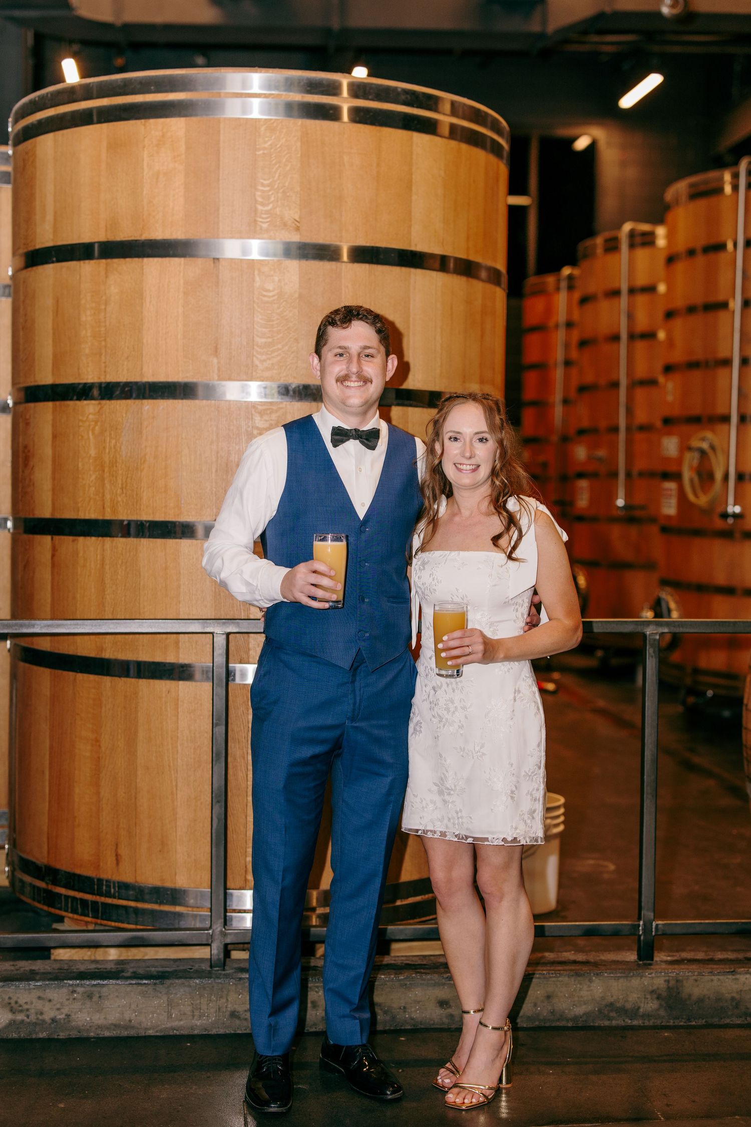 Couple in wedding attire holding beer glasses in front of large wooden barrels at a brewery venue.