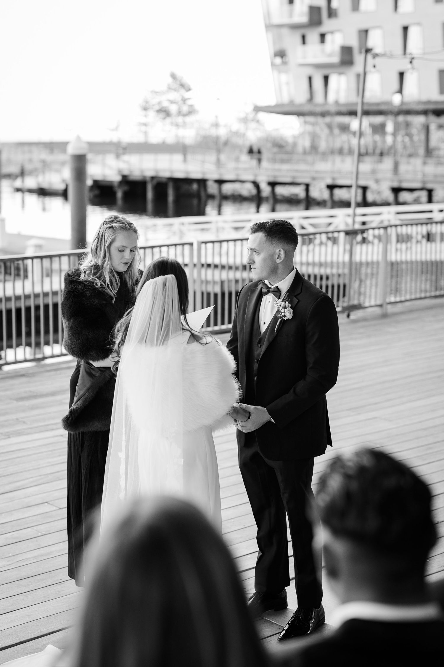 Black and white photo of a bride and groom exchanging vows outdoors on a waterfront deck with an officiant.