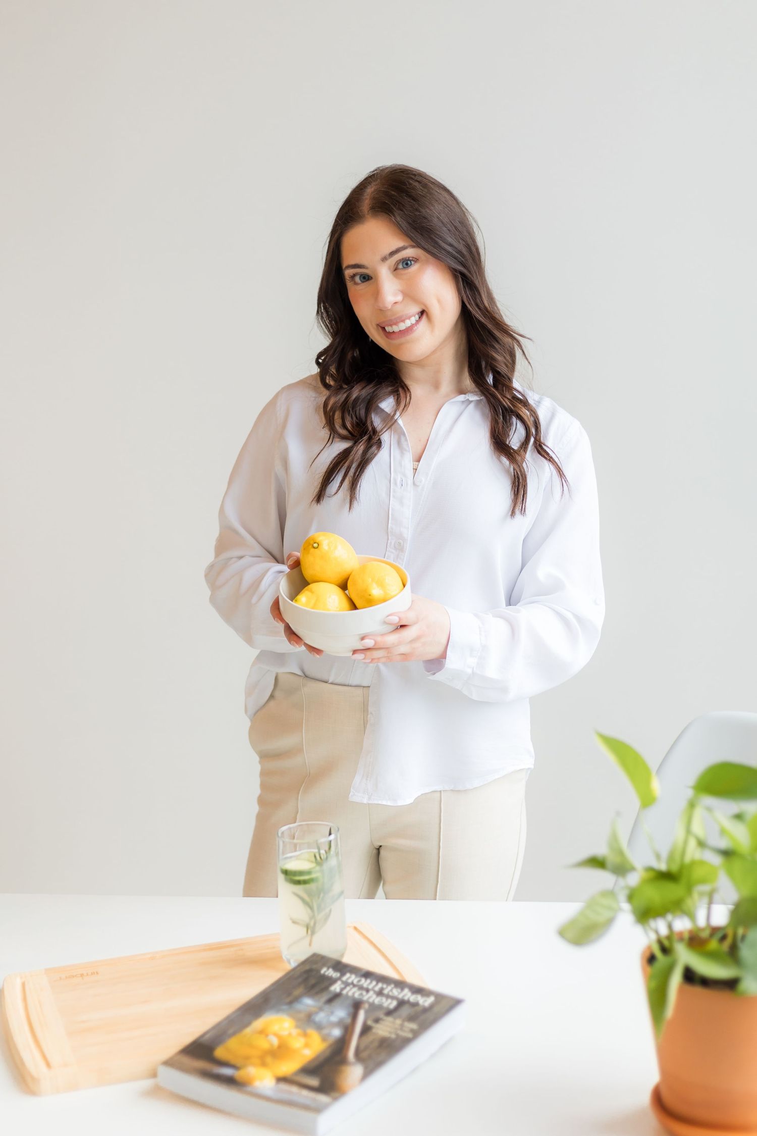 Smiling woman in white shirt holding bowl of lemons near table with cookbook and plant.