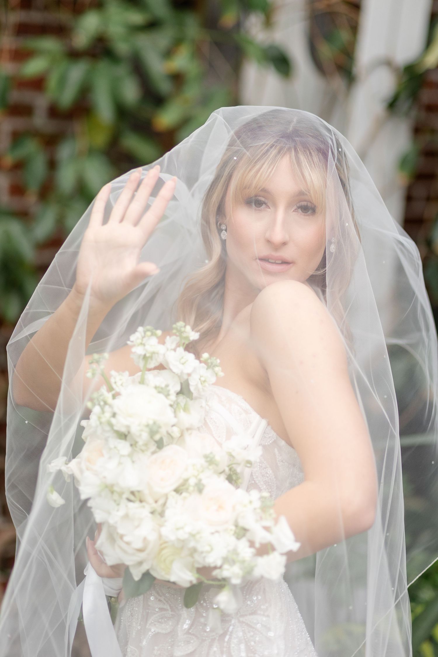 Bride under veil holds white bouquet and raises hand toward camera outdoors.