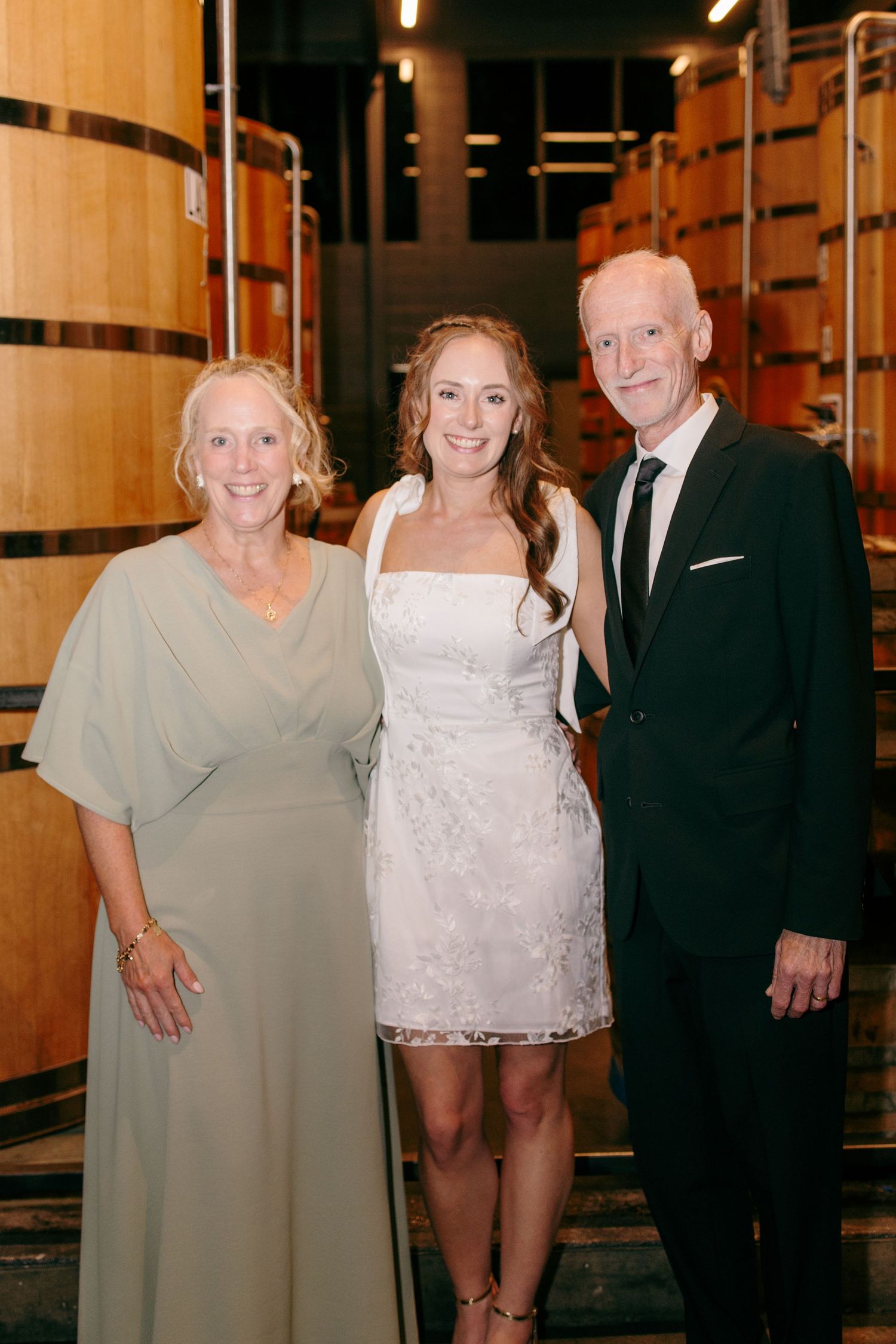Bride in short white dress posing with parents in front of wooden barrels at brewery wedding.