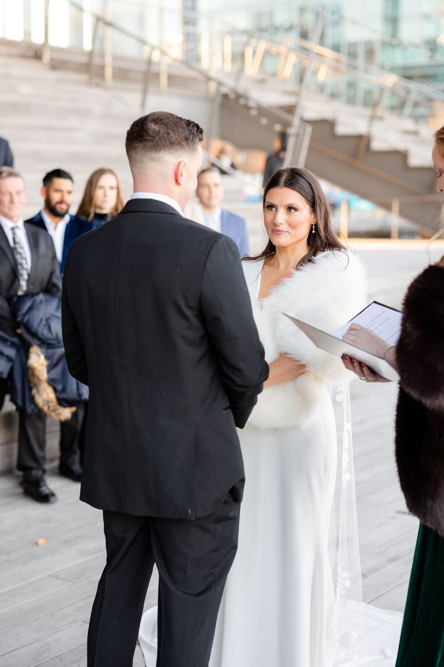 Bride in white gown and fur wrap exchanges vows with groom in black suit at outdoor winter wedding ceremony.