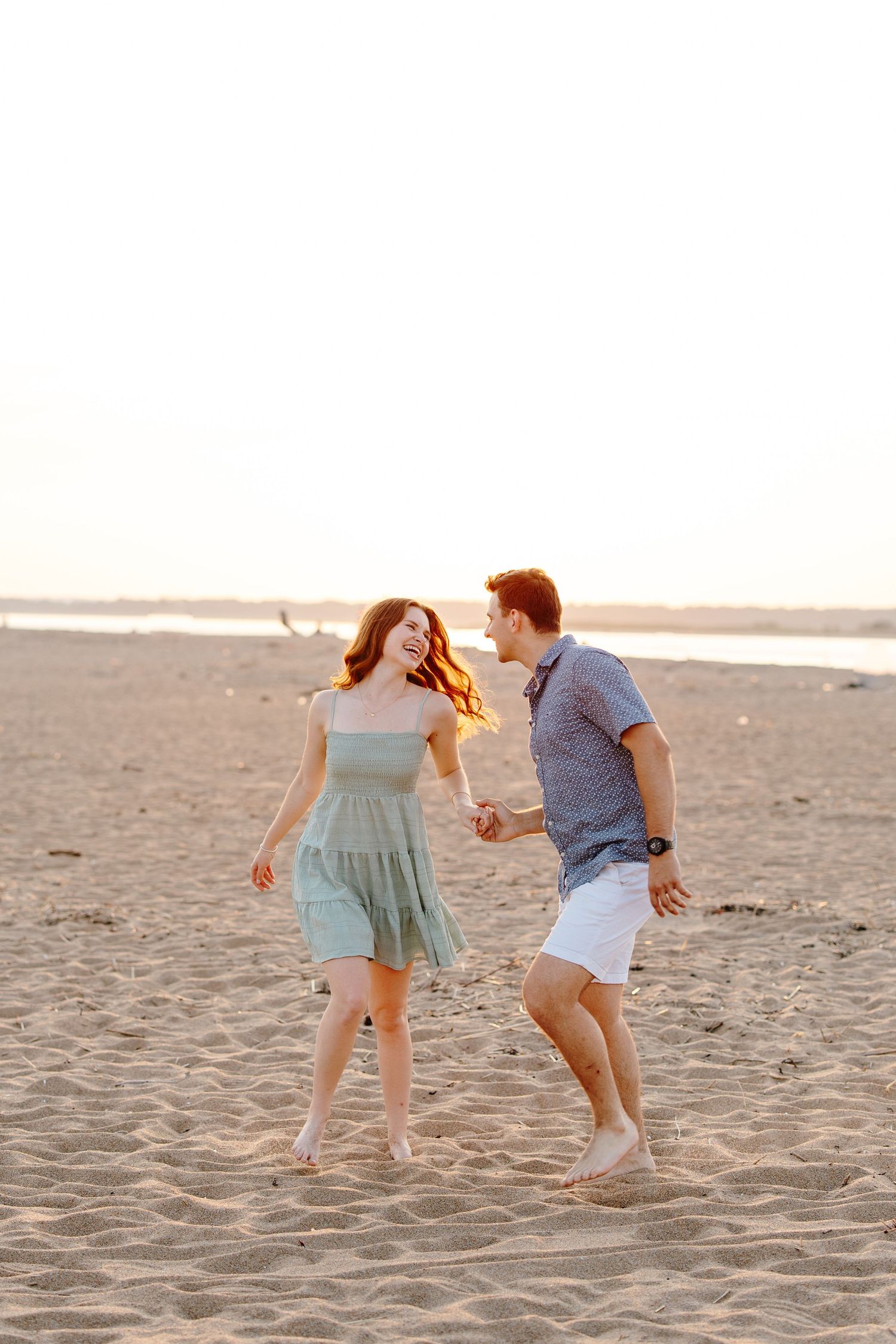 A joyful couple holds hands and dances on a sandy beach at golden hour, the woman in a mint dress laughing.