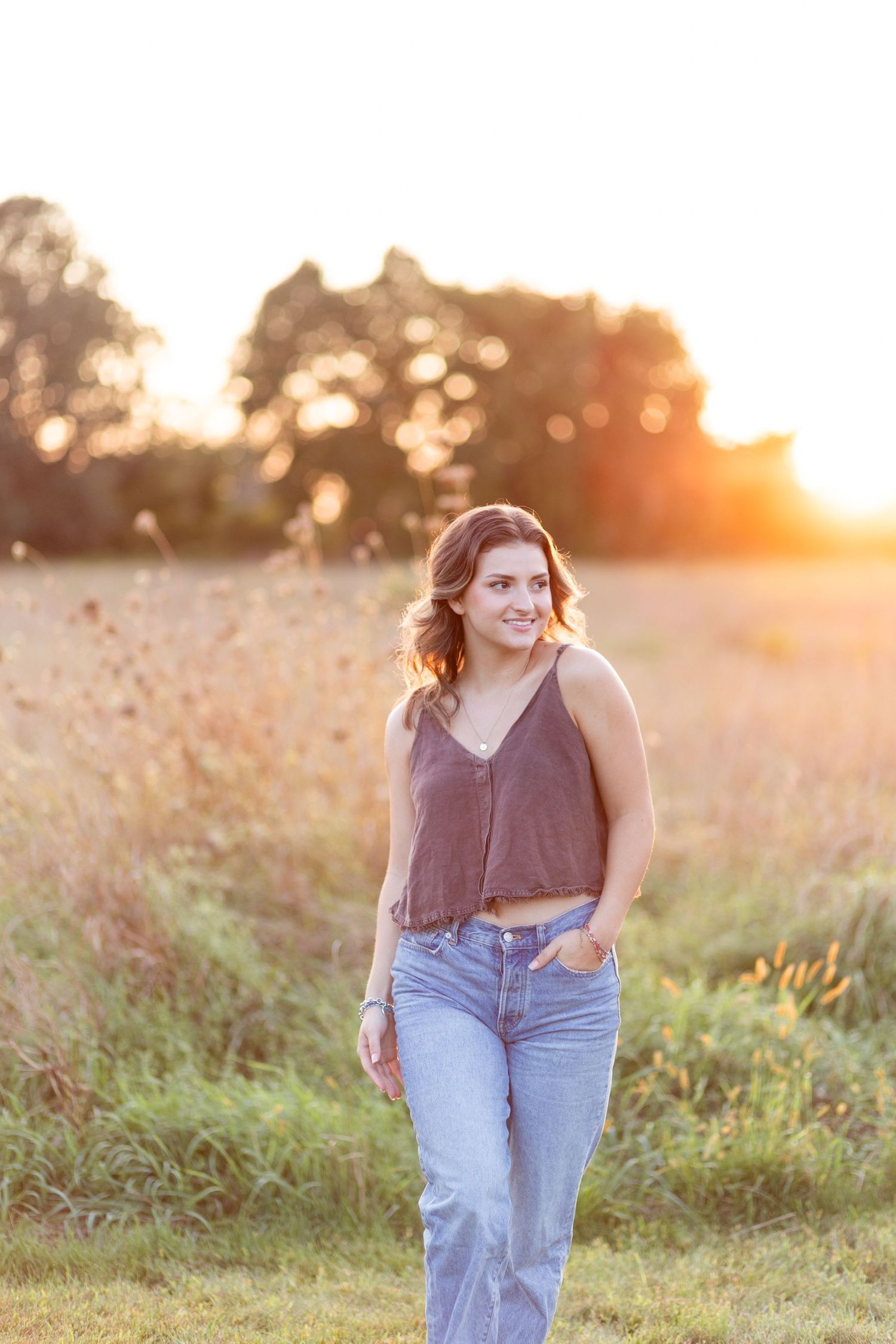 Smiling young woman in gray crop top and jeans walking through golden sunlit meadow at sunset.