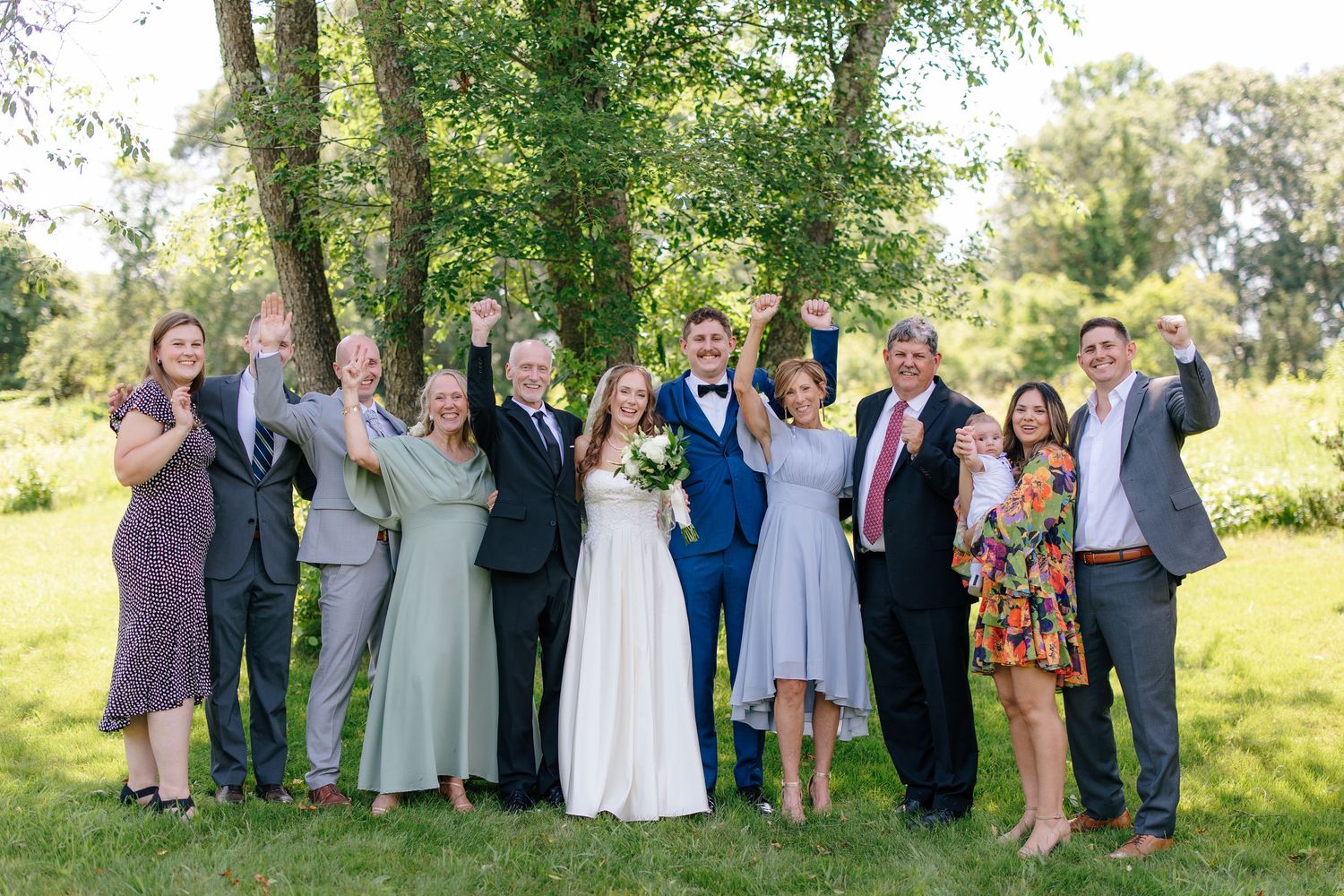 Wedding party and family celebrating outdoors with raised hands among lush green trees.