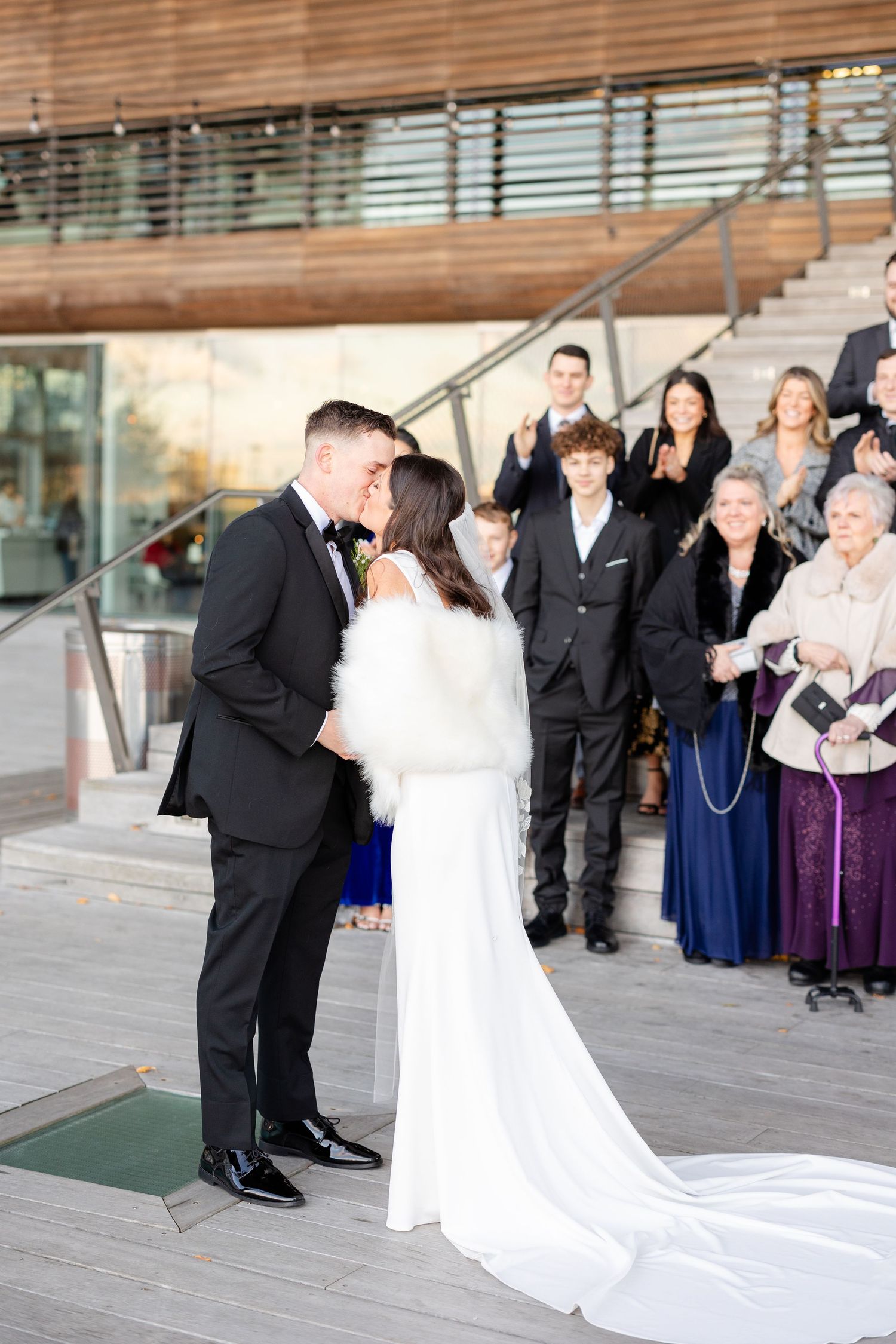 Bride and groom share first kiss at outdoor wedding ceremony with guests applauding.