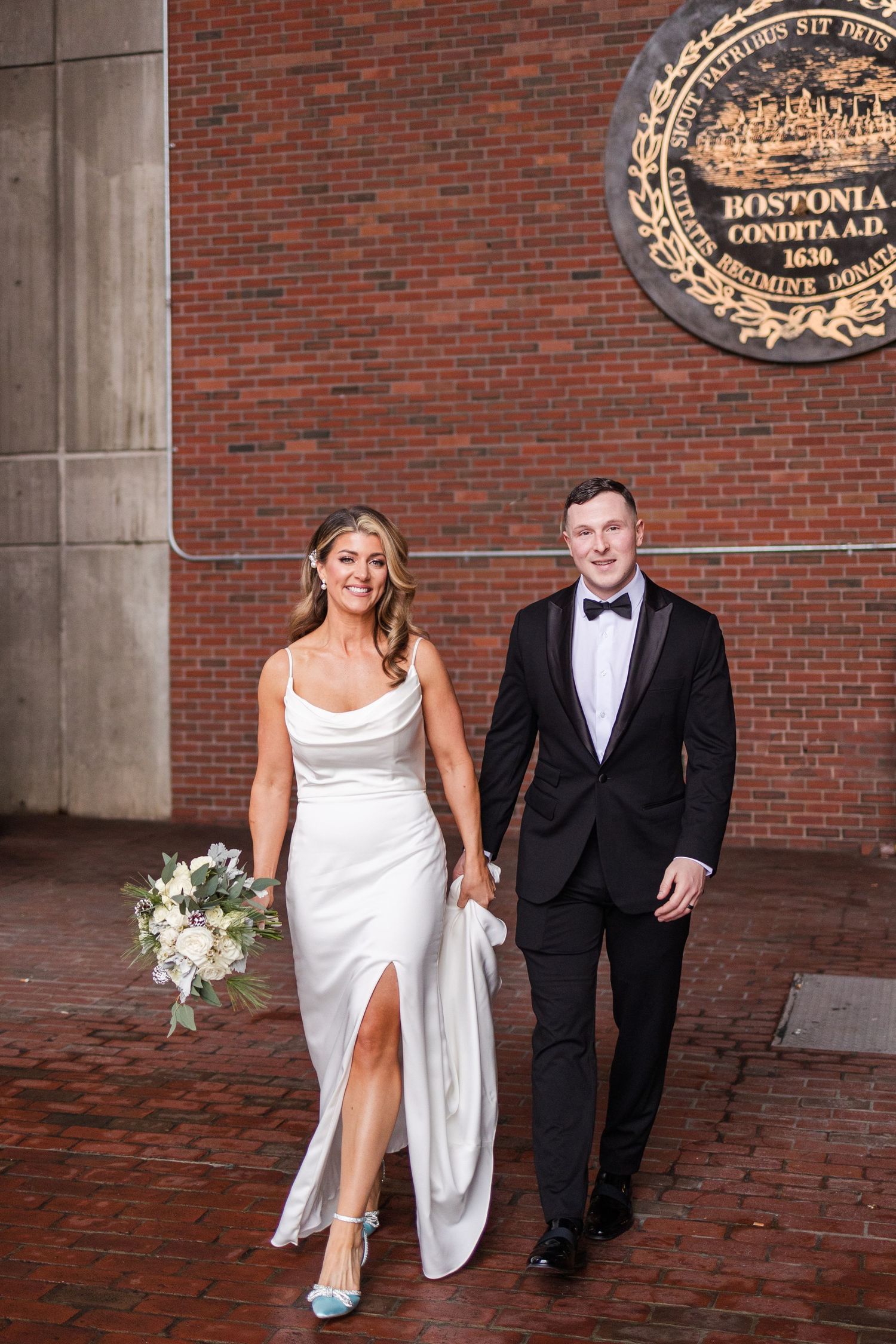 Bride and groom walking hand in hand near the historic Boston City Hall seal on brick plaza.