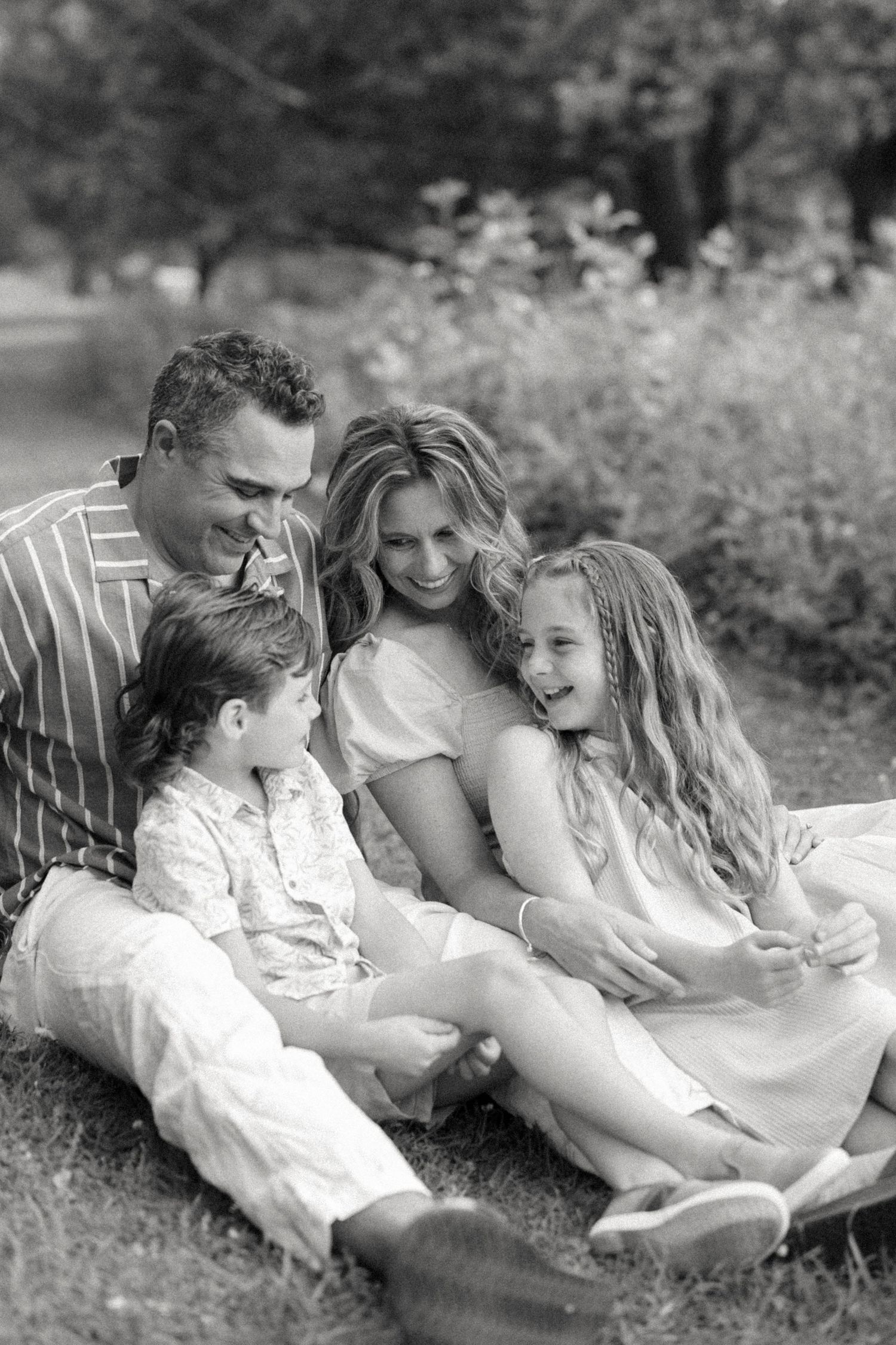 A black and white photo of a happy family of four laughing together while sitting on grass in a park.