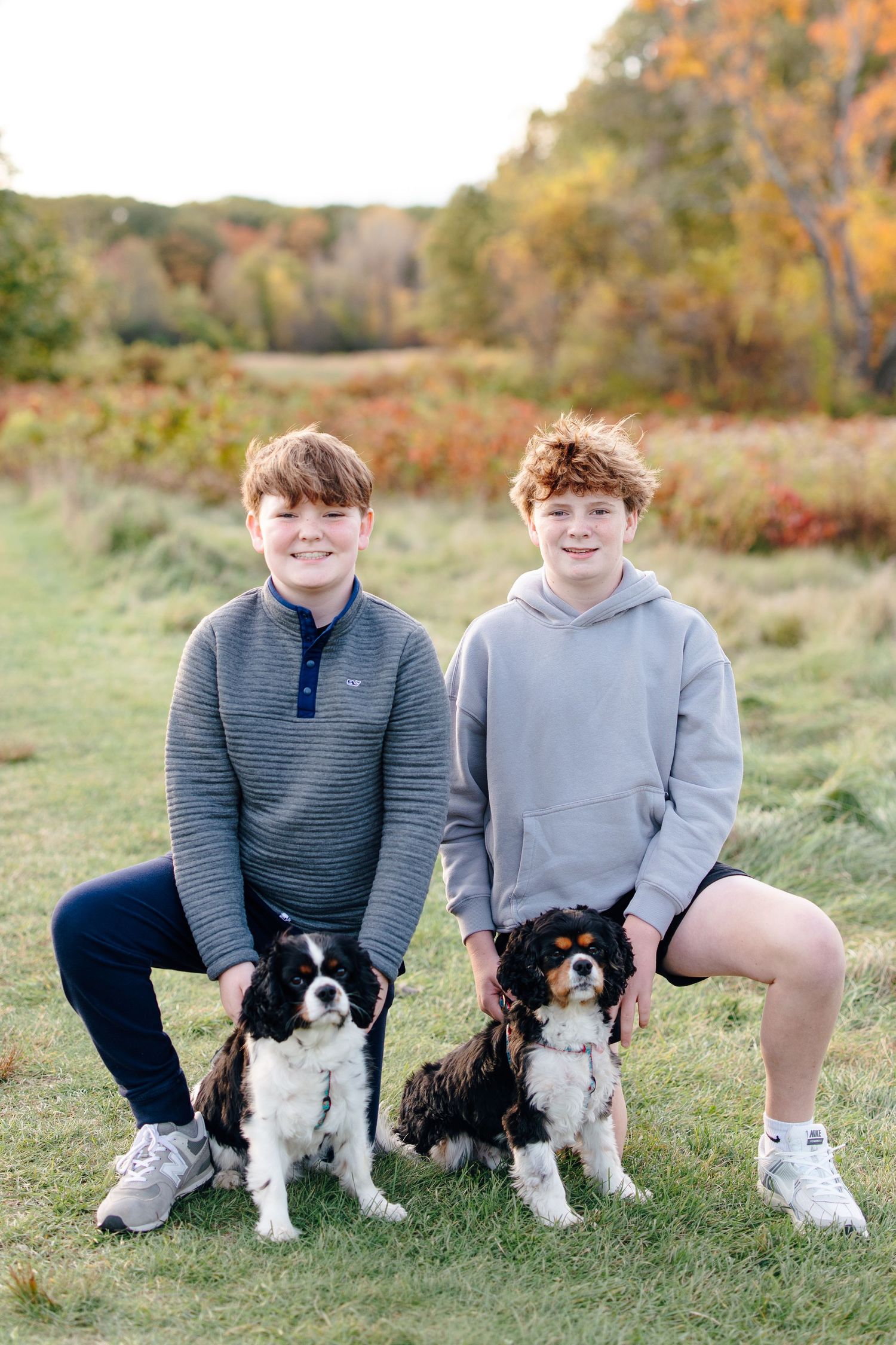 Two smiling teenage boys pose outdoors in a fall field with their two Cavalier King Charles Spaniel dogs.
