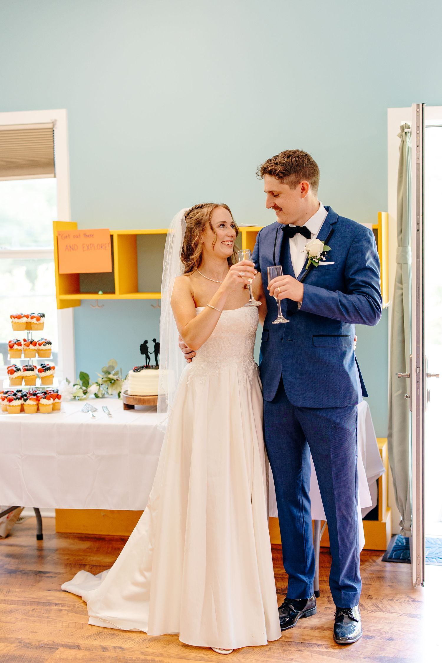 Bride and groom smile while toasting champagne glasses near wedding cake table in blue-walled reception venue.
