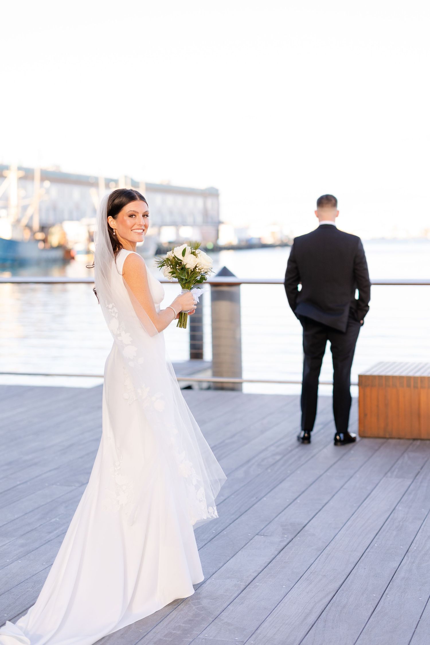 Smiling bride in white gown holds bouquet while groom stands facing waterfront harbor in background.