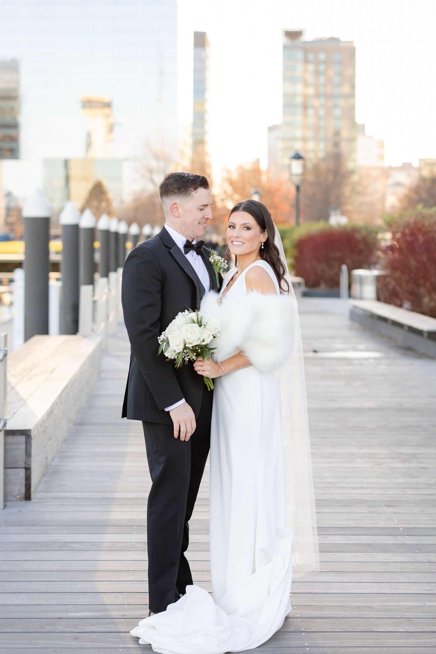 Bride and groom pose on a waterfront boardwalk, she in a white gown with fur wrap holding bouquet, he in a black tuxedo.