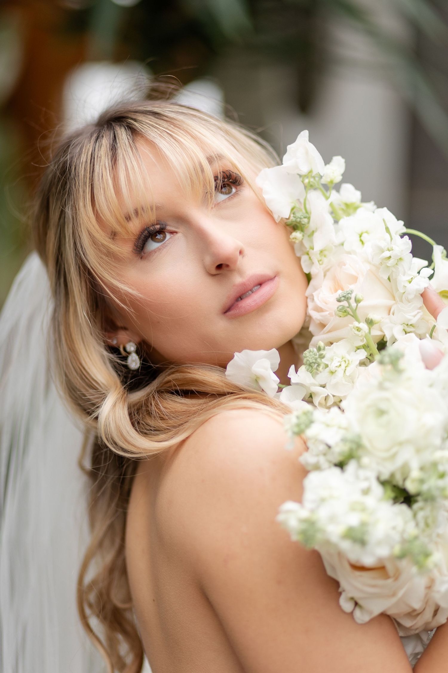 Blonde bride with bangs holds white floral bouquet close to her face.