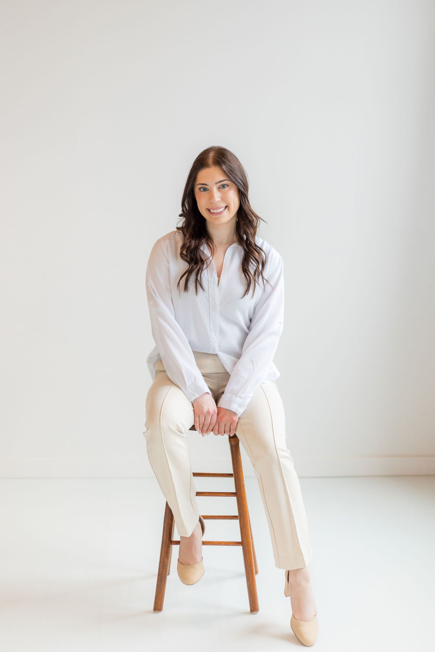 Young woman in white blouse and beige pants sitting on wooden stool smiling against white background.