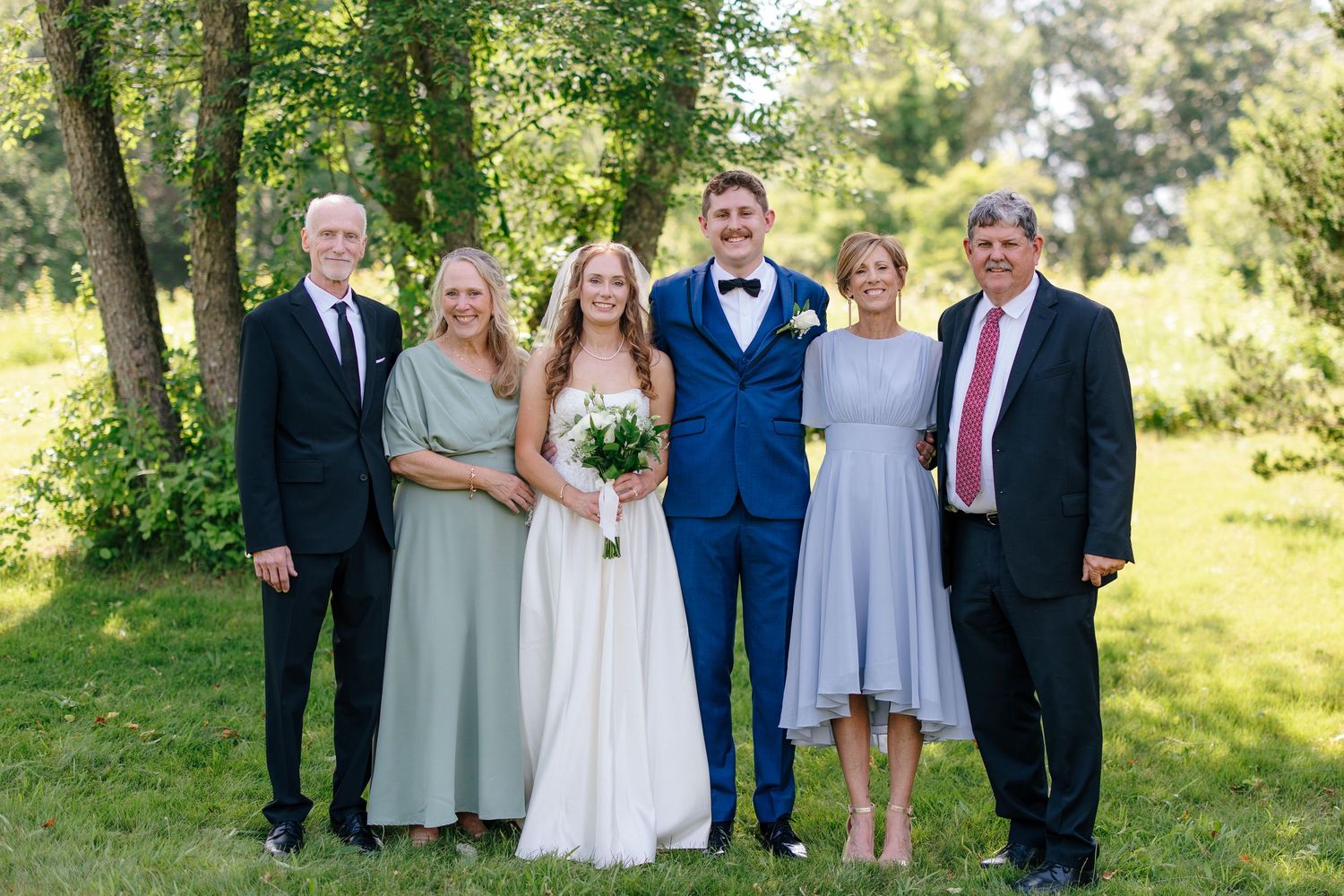 Bride and groom posing with family members in formal attire on green lawn with trees.
