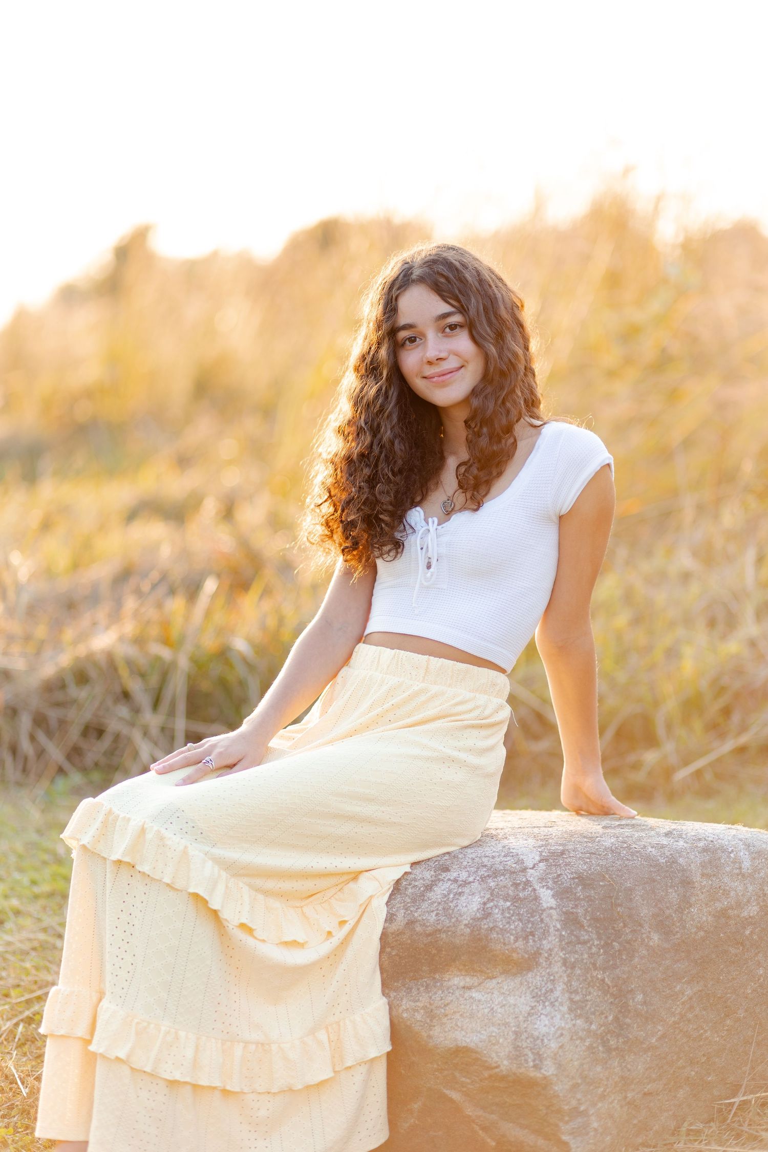 Teenage girl with curly hair in white crop top and yellow maxi skirt sitting on rock at sunset.