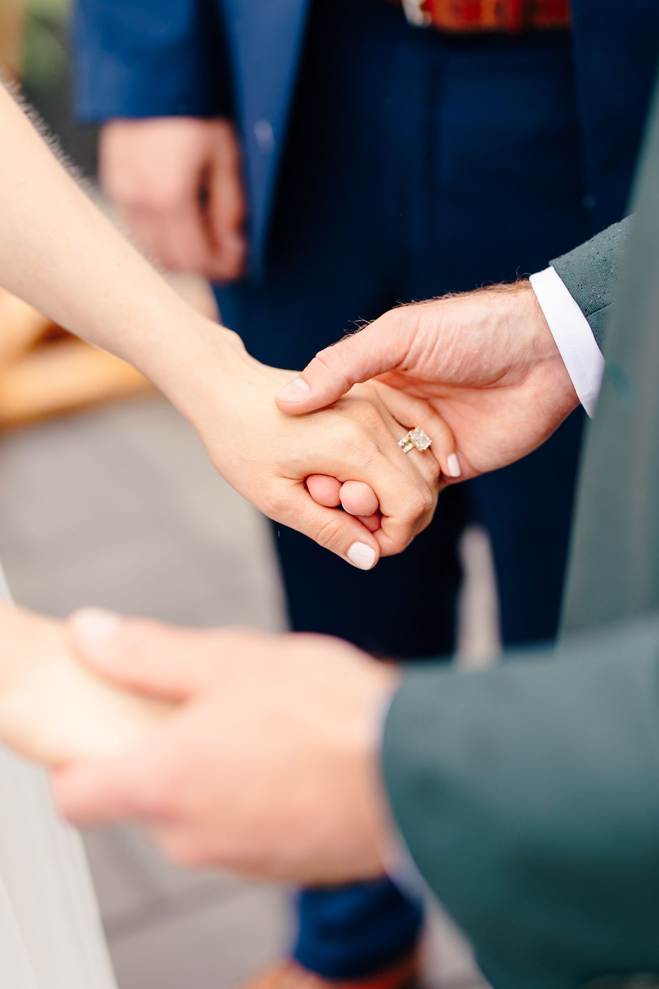 Couple holding hands during wedding ceremony, bride wearing diamond engagement ring, groom in gray suit.