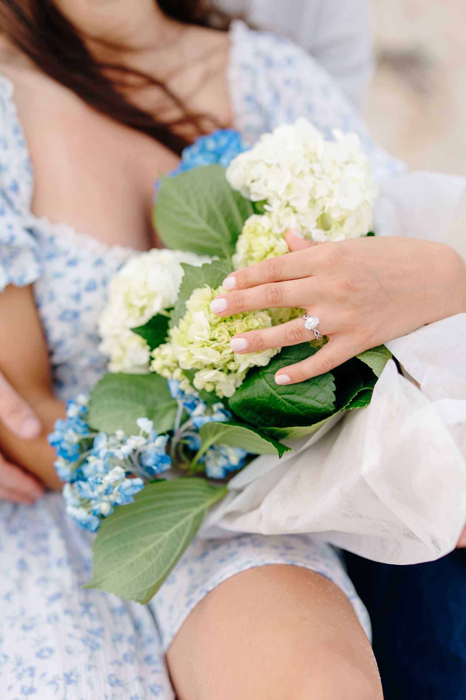 Close-up of engaged couple holding blue and white hydrangea bouquet with diamond ring visible.
