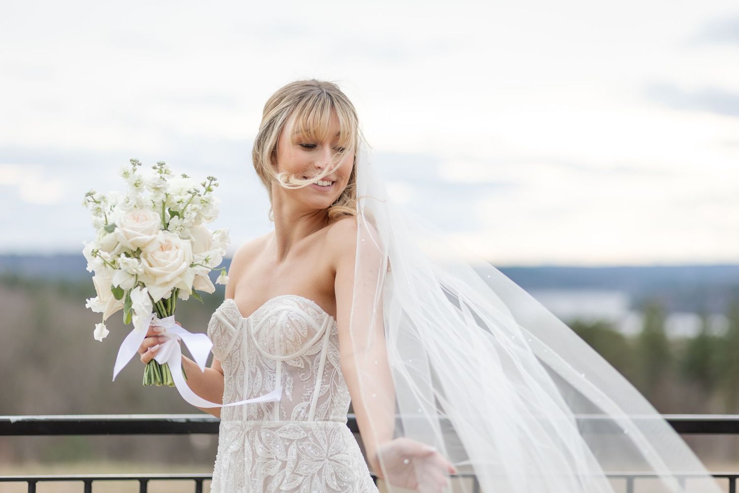 Bride in strapless beaded gown holds white bouquet on outdoor balcony with veil.