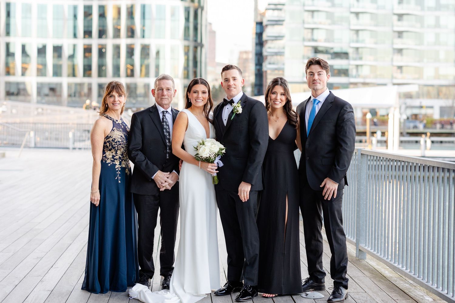 Wedding party of six posing on waterfront boardwalk with city skyline backdrop.
