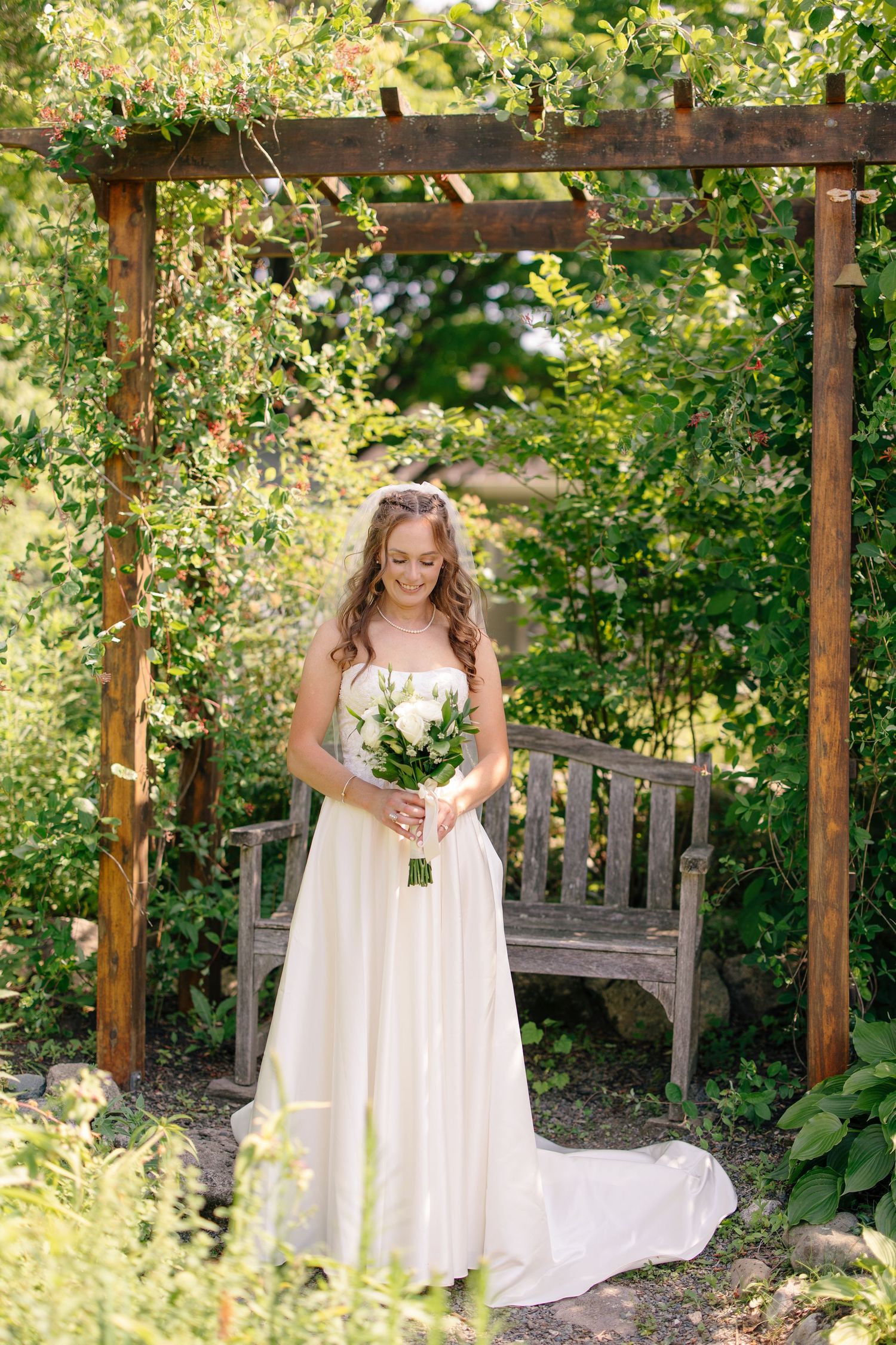 Bride in white gown holding bouquet standing under wooden pergola surrounded by green garden.