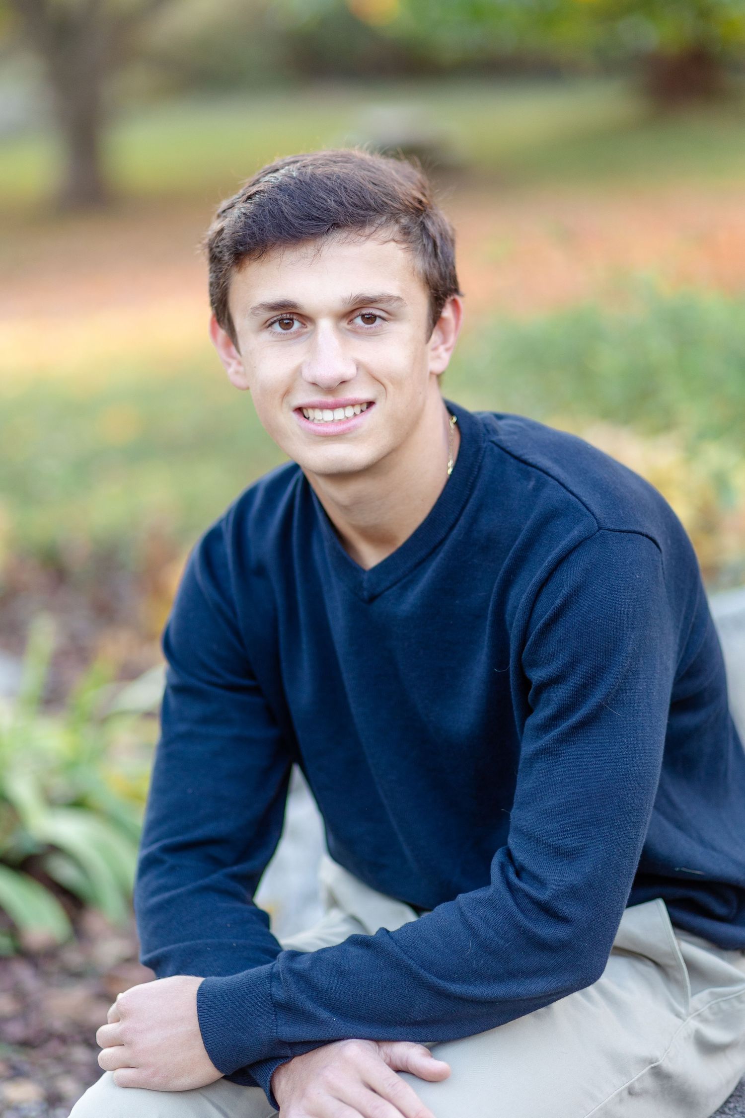 A smiling teenage boy in a navy sweater crouching outdoors in a garden with autumn foliage in the background.