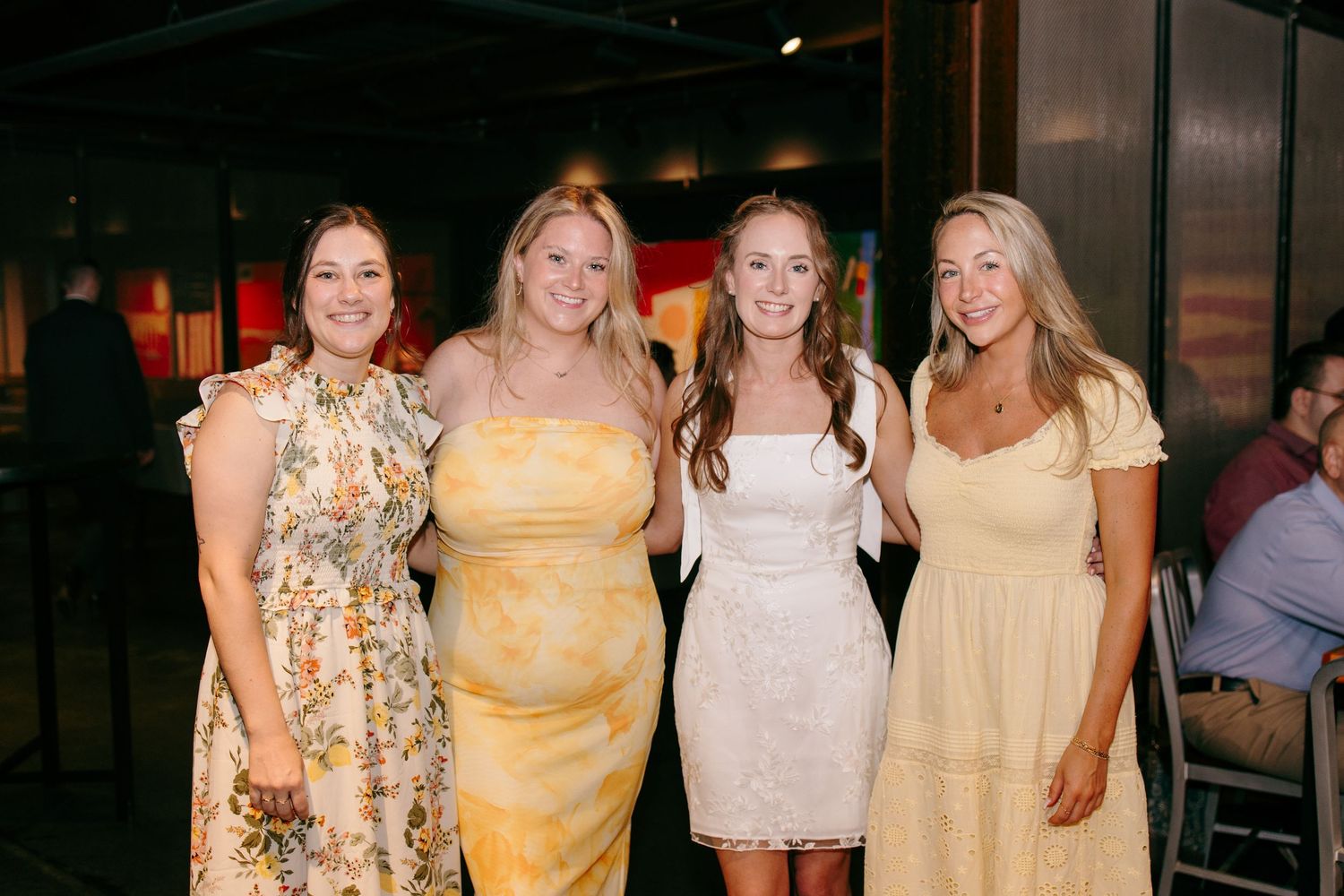 Four women in yellow and white dresses smiling together at a wedding reception celebration.